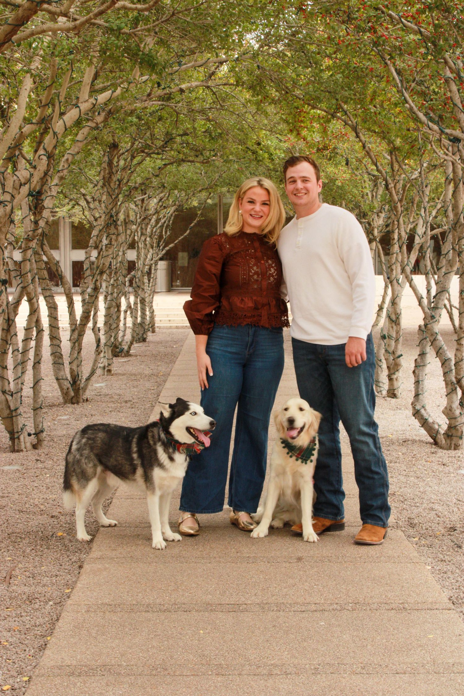 A couple poses with their two husky dogs on a tree-lined pathway.