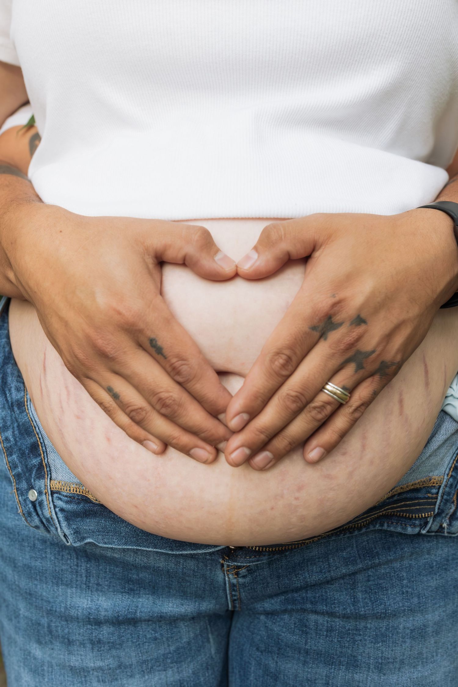 Two hands forming heart shape on pregnant belly, one with tattoos and gold ring, over white shirt and jeans.