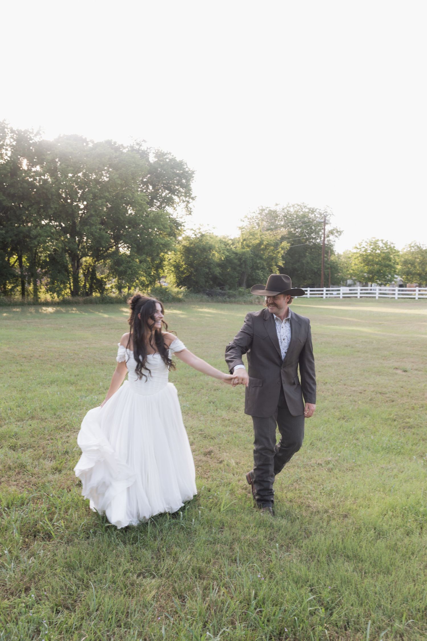 Bride in flowing white lace gown holds hands with groom in cowboy hat as they walk through a sunny green field.