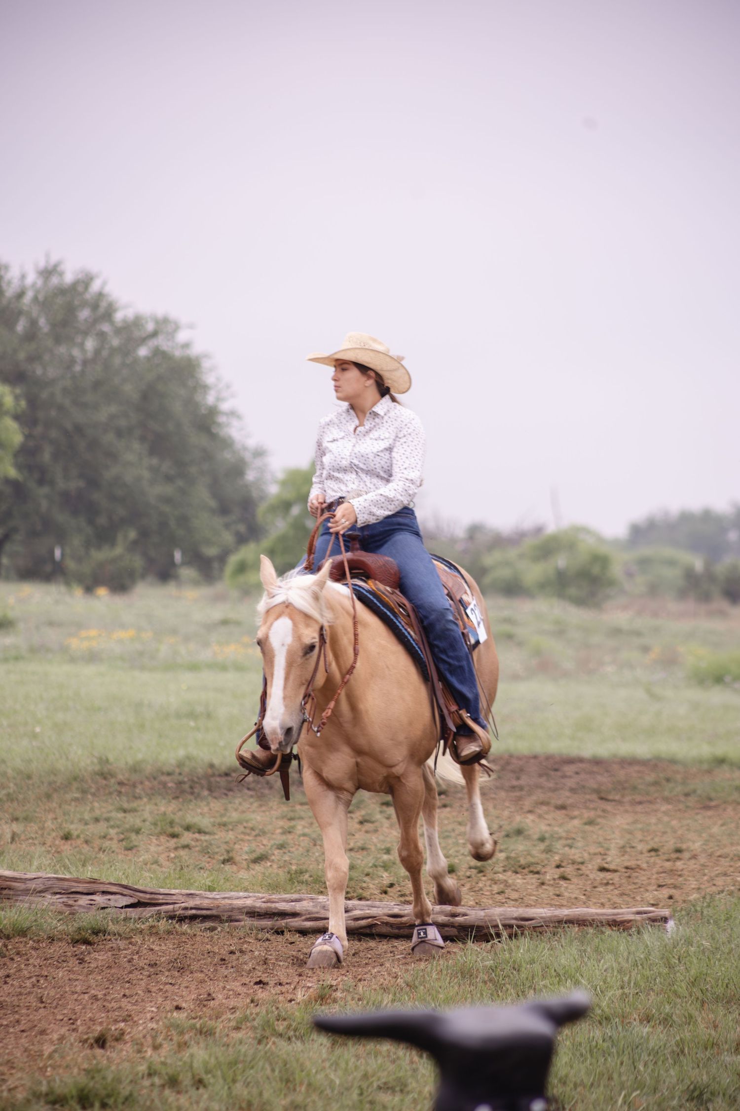 A cowgirl in a straw hat rides a palomino horse across a green ranch field.