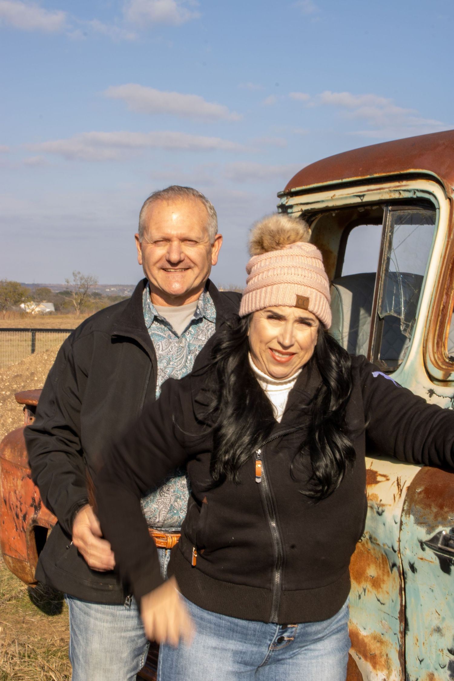 Two people pose playfully in front of a rustic vintage truck in a rural setting on a sunny day.
