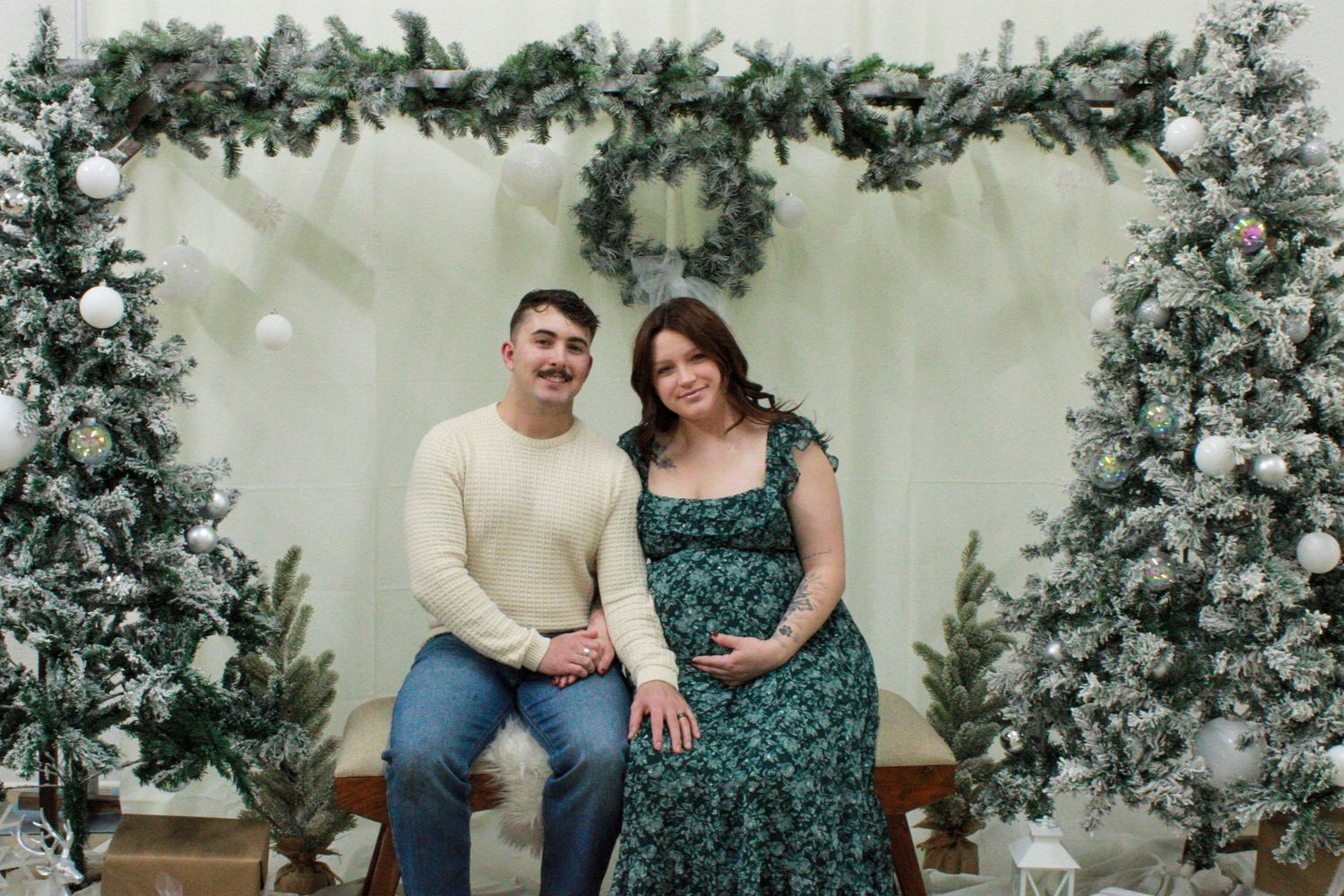 A couple sits together on a wooden bench surrounded by lush greenery and floral decorations against a light wall.