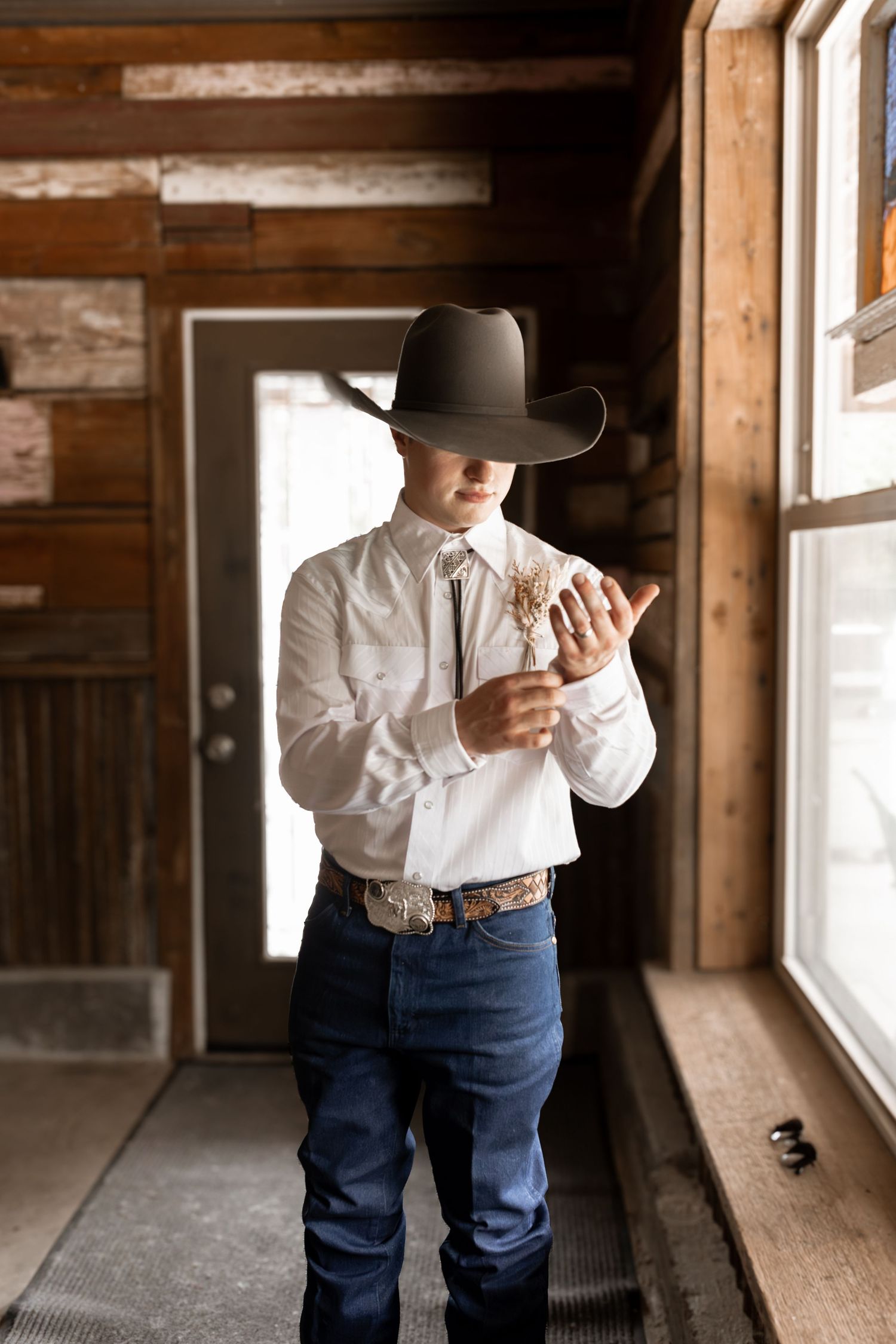 Man in cowboy hat, white shirt, and jeans adjusting cufflinks in a rustic wooden interior.