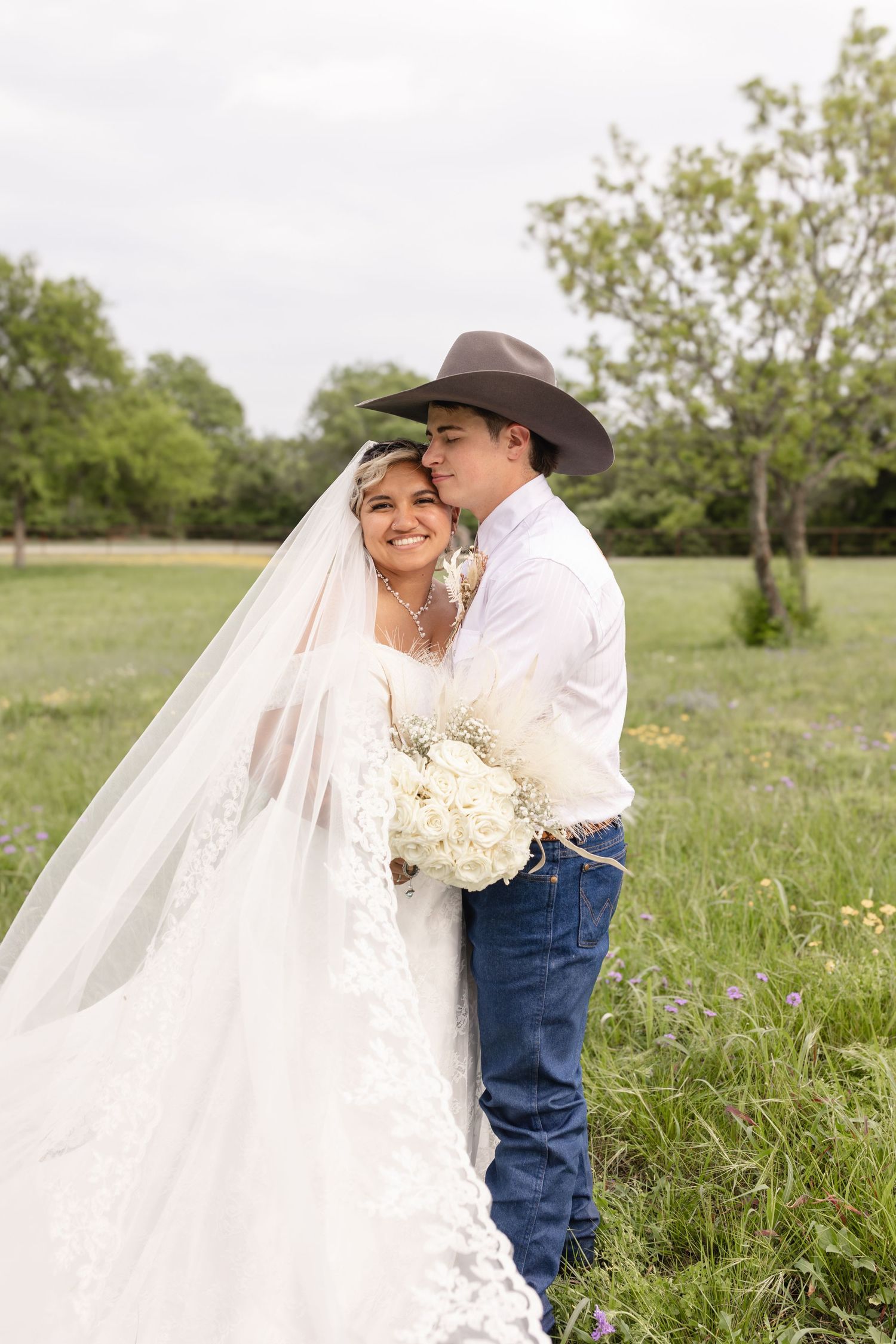 Bride and groom pose in a green field, she holds white flowers, he wears a cowboy hat and jeans.