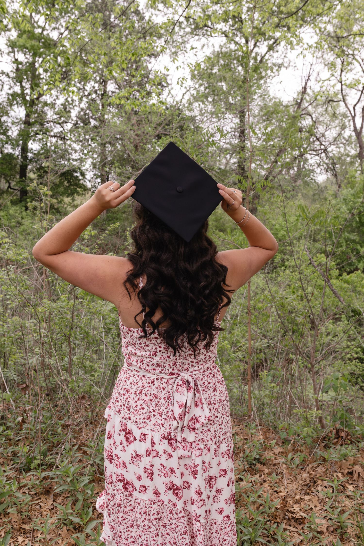 A young woman in a floral dress holds a black graduation cap on her head while standing in a lush green forest.