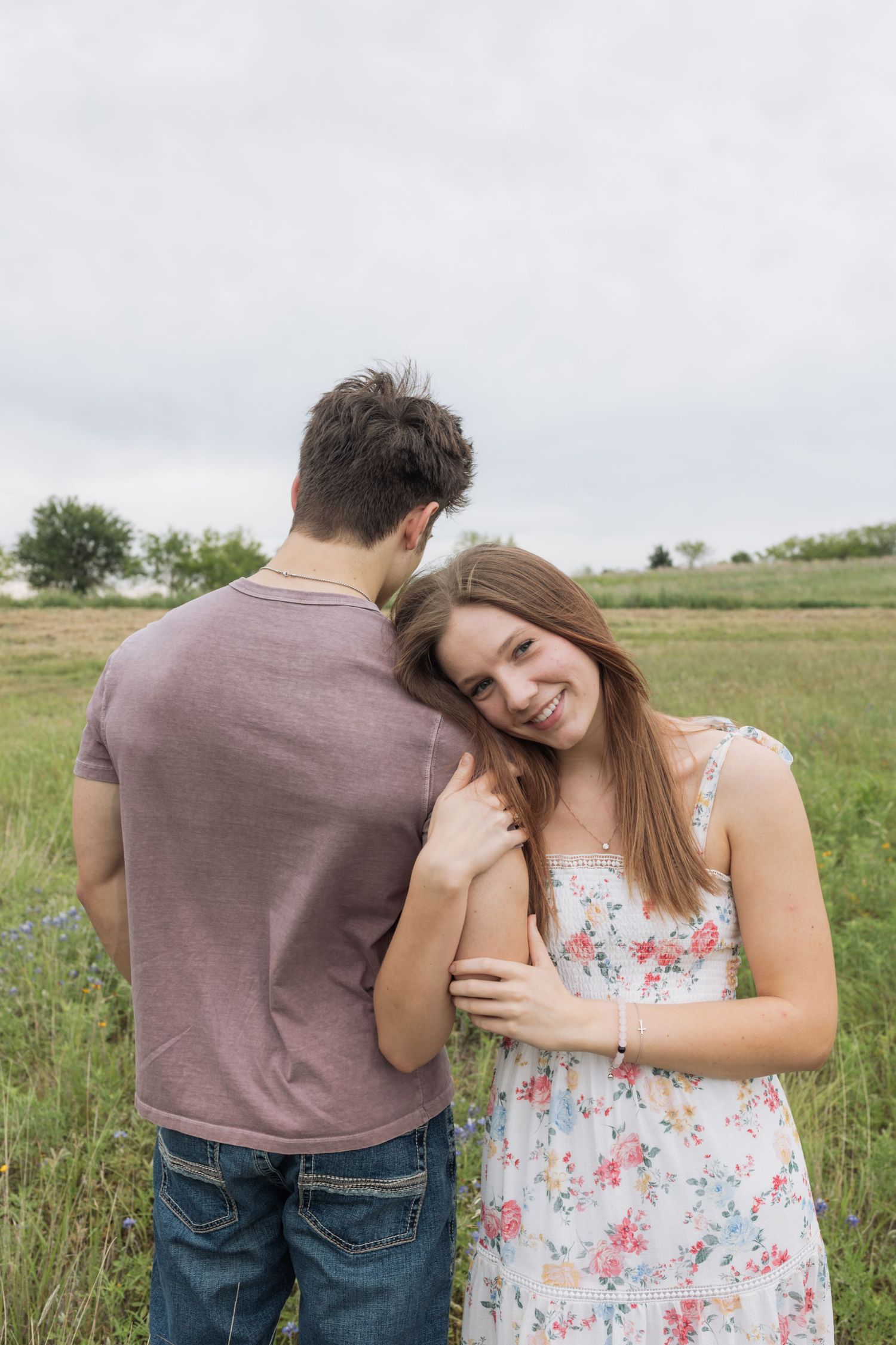 Smiling young woman in floral dress leans on man's shoulder in green field.