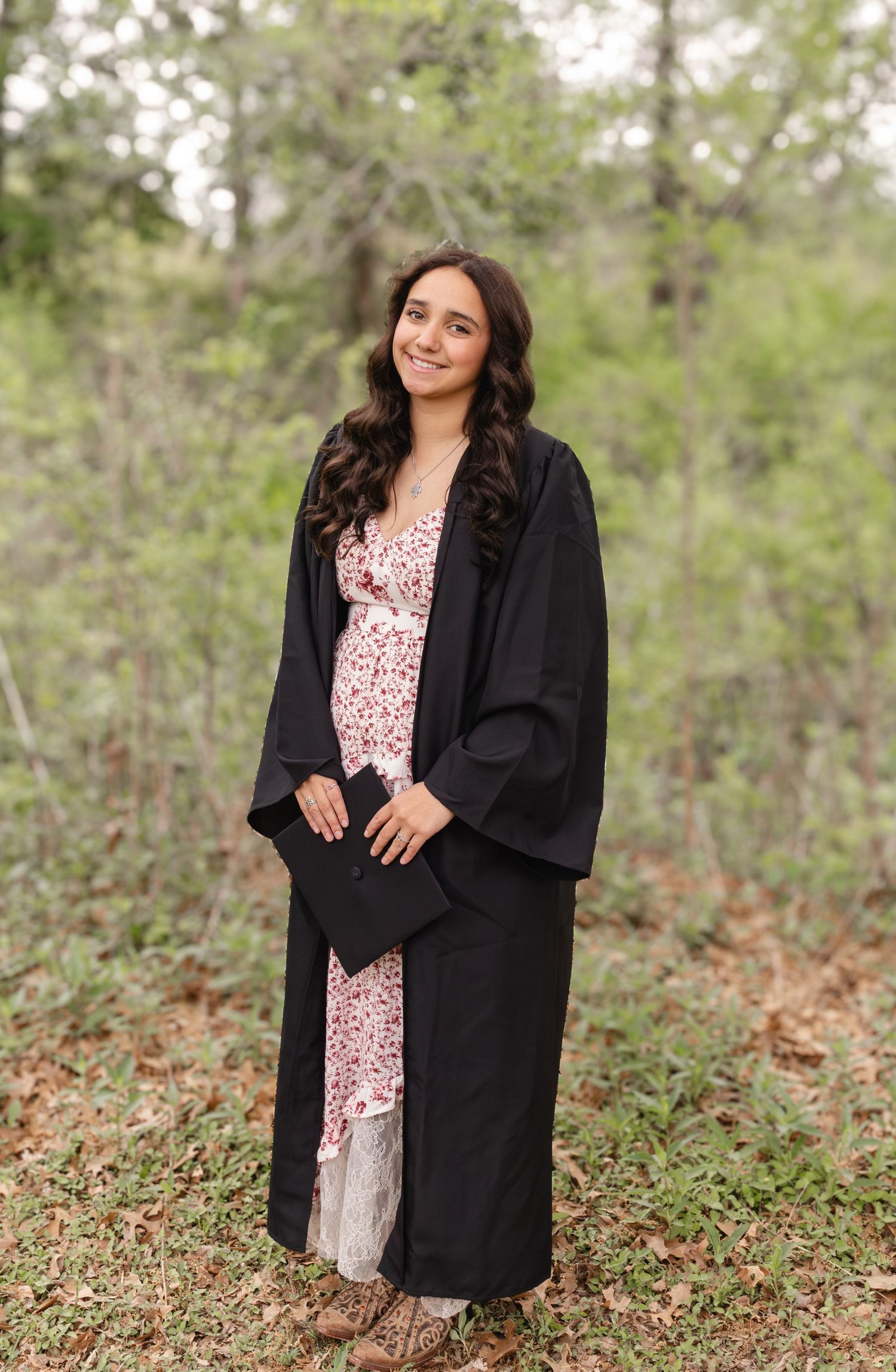 A person poses outdoors in a black coat and floral dress against a wooded natural background.