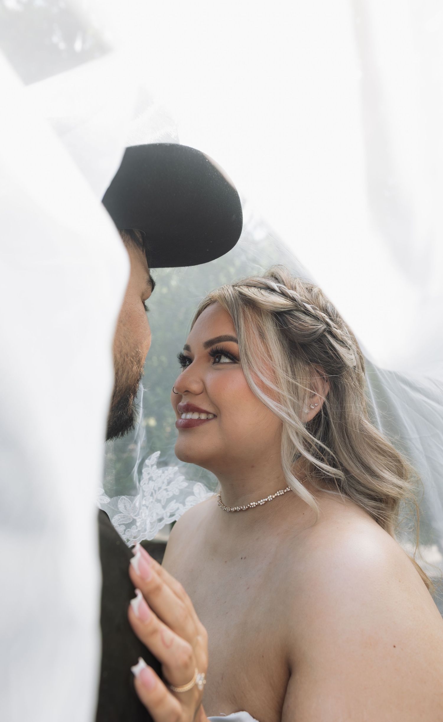 Bride smiling up at groom under flowing white veil, wearing diamond choker necklace and braided updo at wedding.