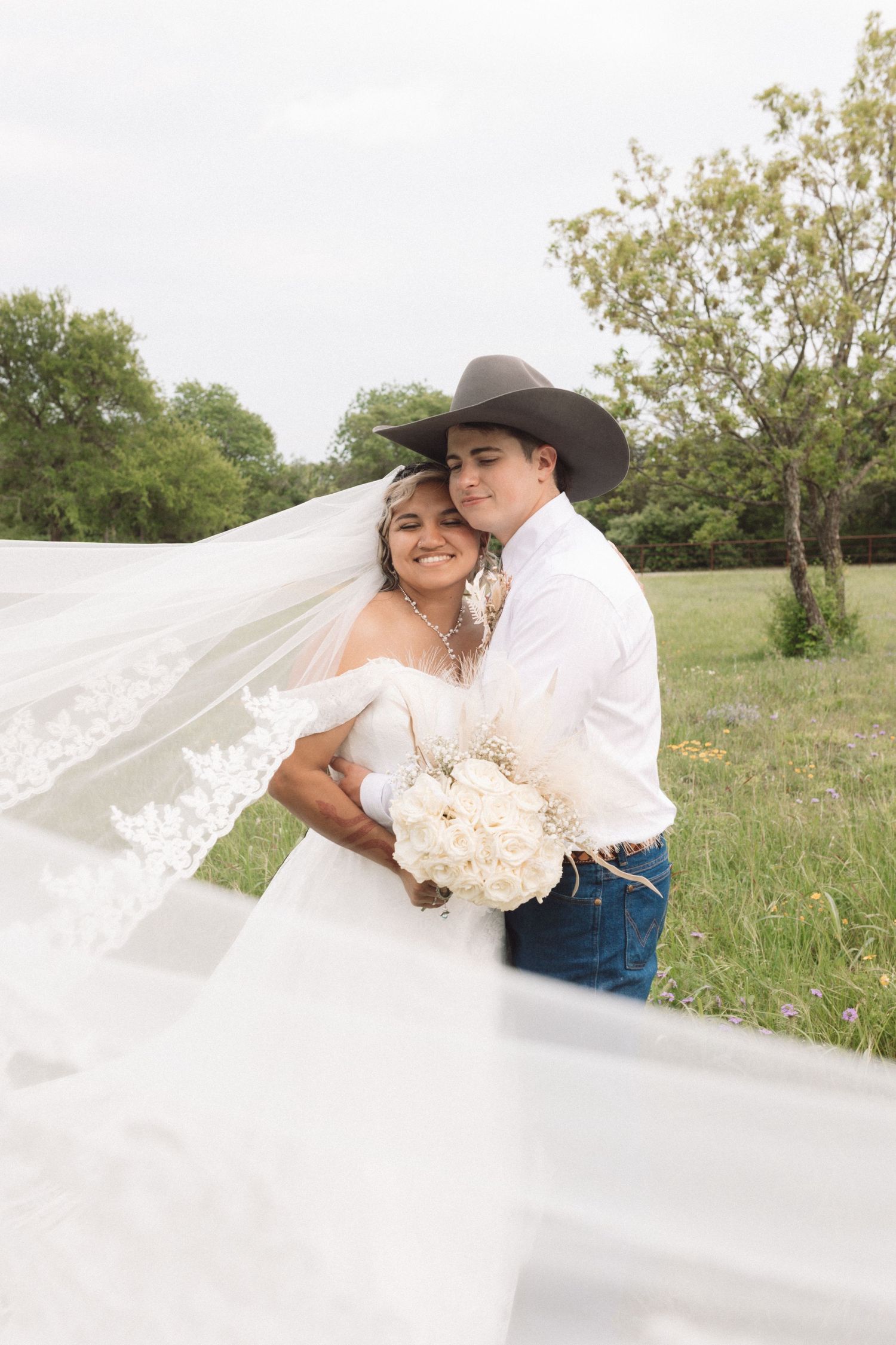 Bride and groom embrace in a field, her lace veil flowing, holding white bouquet, groom in cowboy hat and jeans.