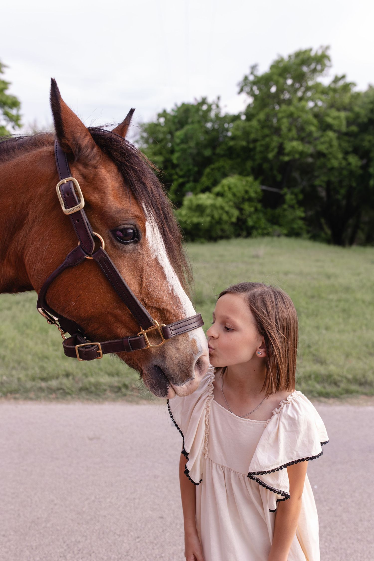 Young girl in white dress kissing a brown horse on the nose outdoors near green trees.