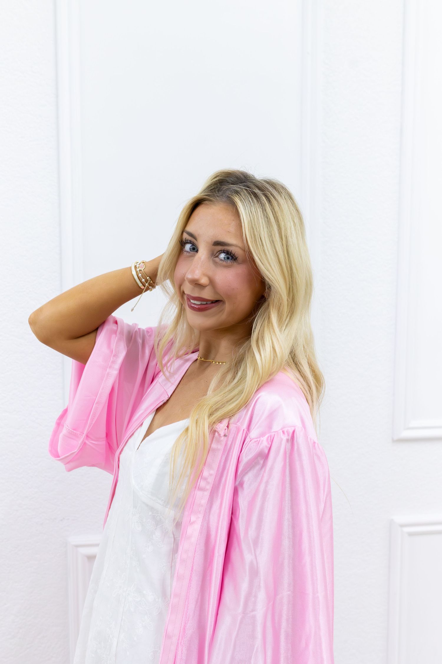 Blonde woman in pink satin shirt and white dress posing against a white door, smiling at camera.