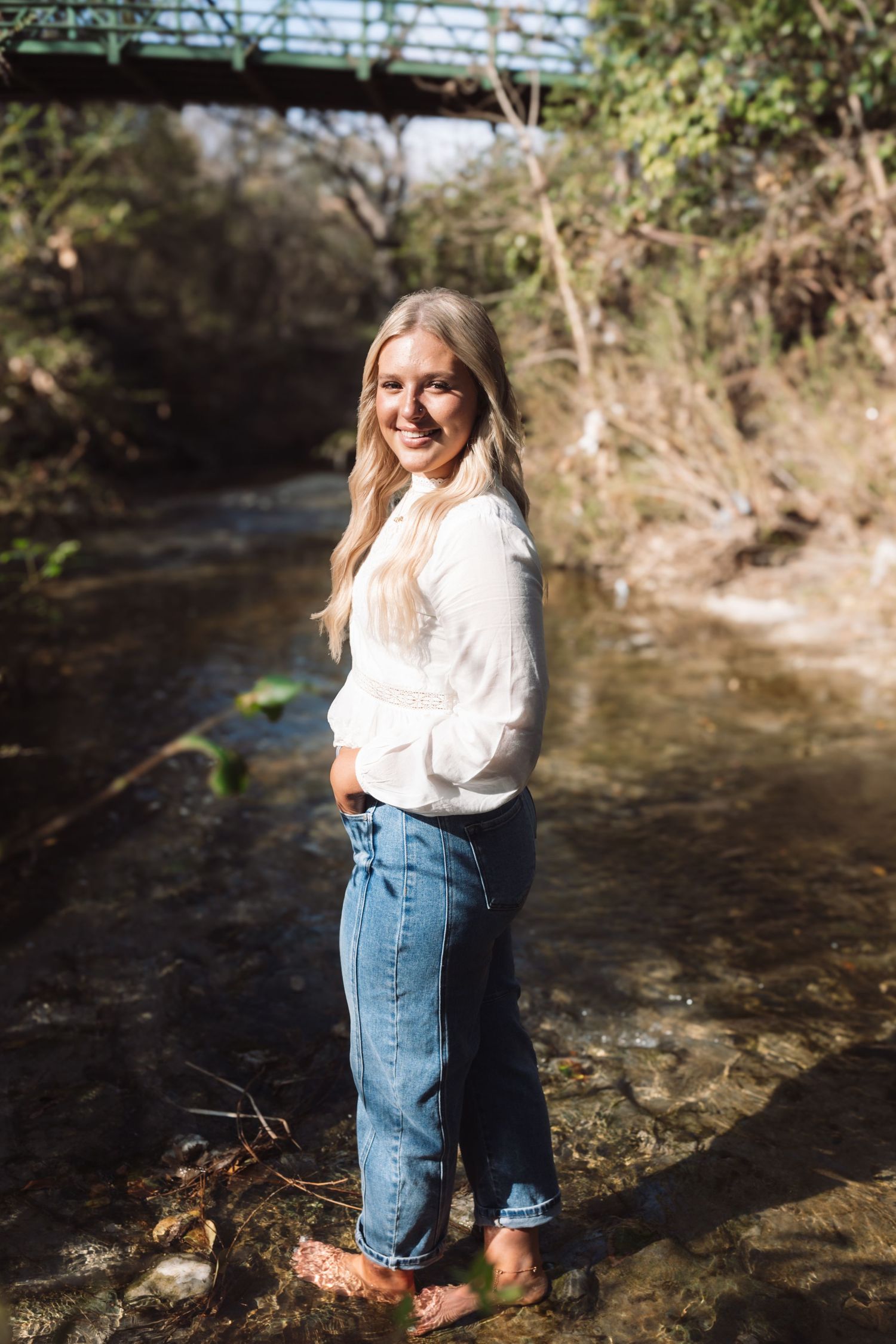 Person in white top and denim skirt stands in shallow creek water on sunny day with natural surroundings.