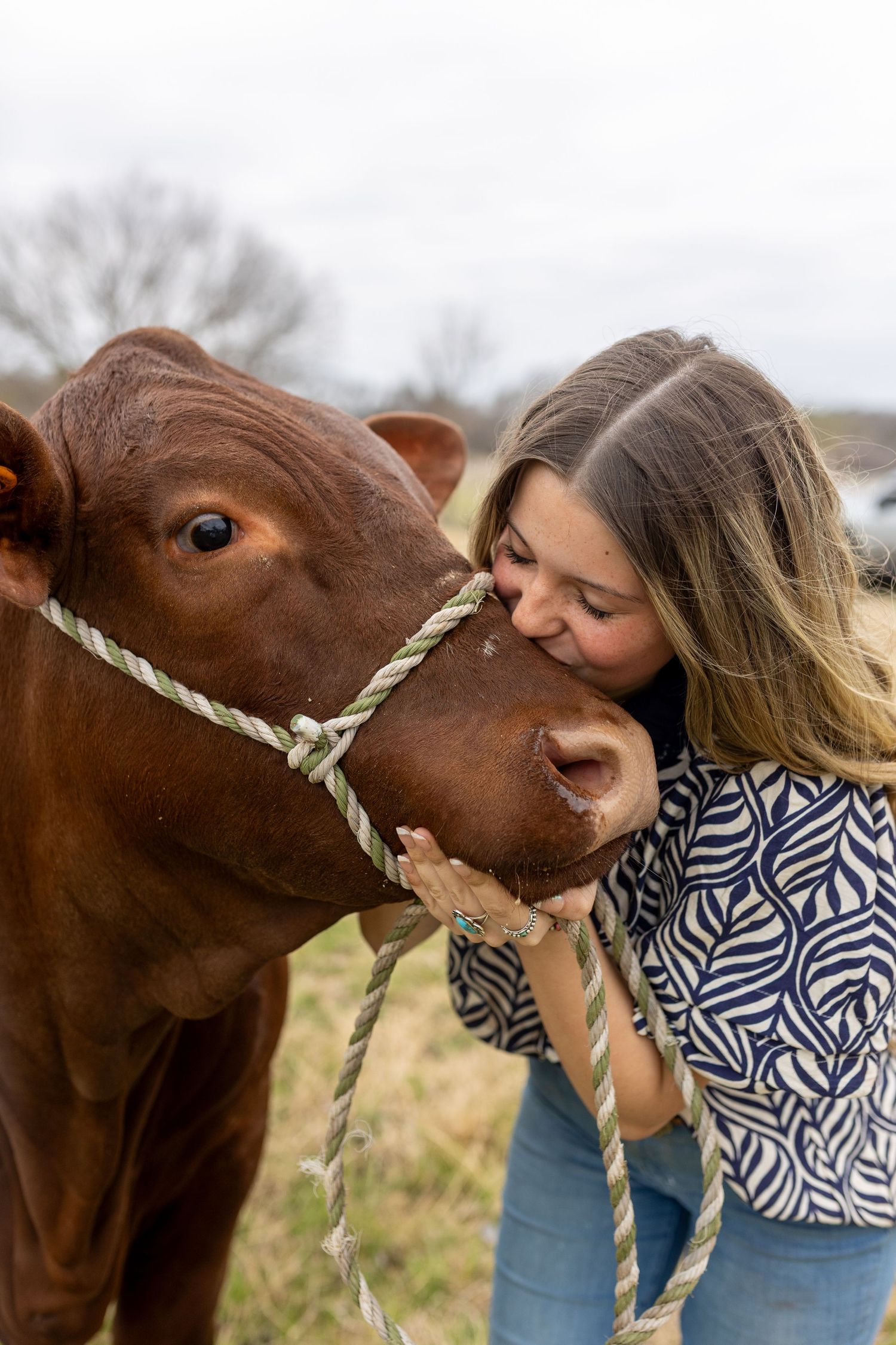A woman in a blue patterned shirt shares a tender moment with a brown horse wearing a rope halter outdoors.
