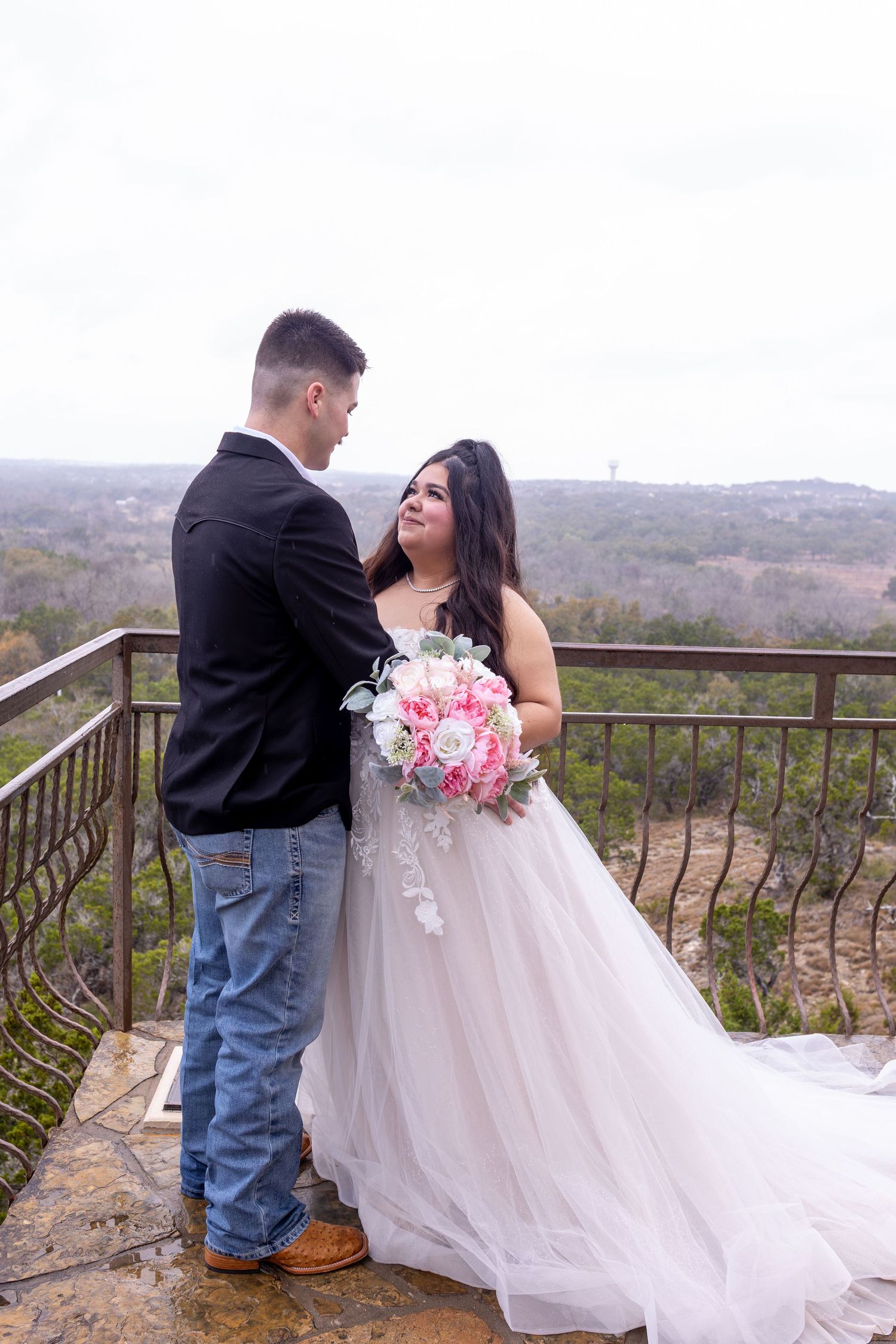 A couple shares a romantic moment on a scenic overlook balcony with the bride holding a pink and white bouquet.