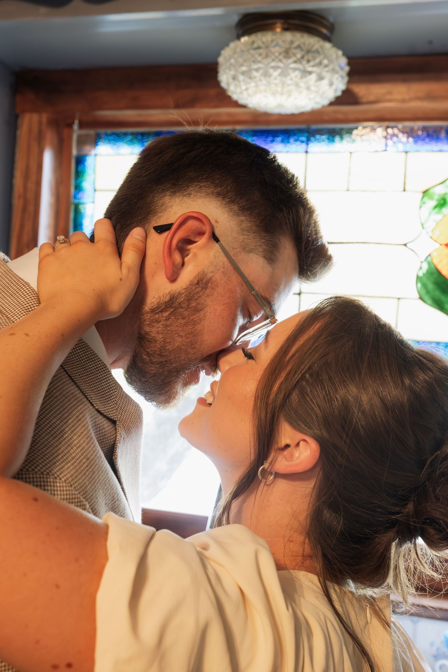 Romantic couple sharing a kiss near a stained glass window, groom wearing glasses and blazer.