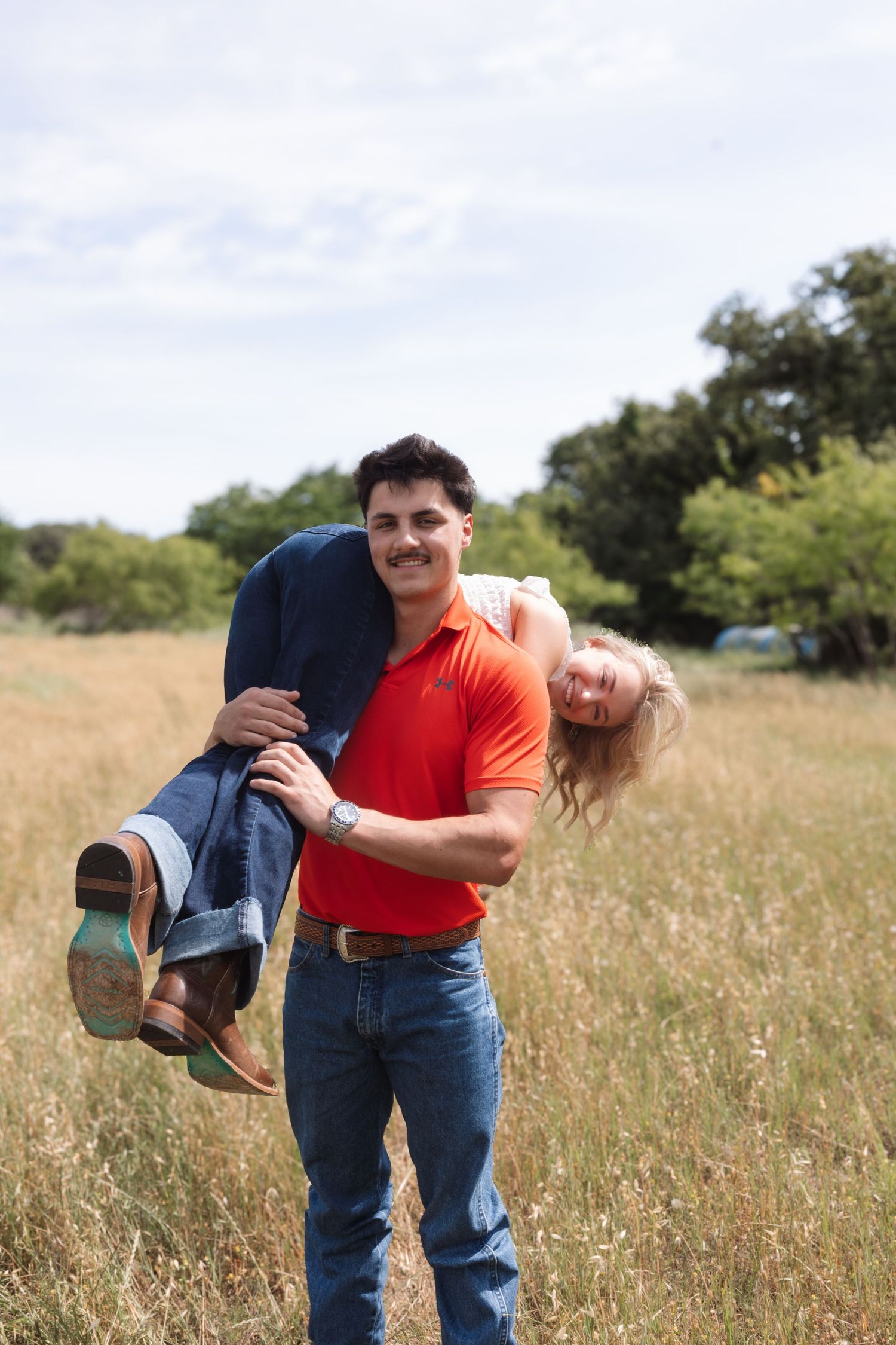 Man in red polo carries smiling blonde woman in sunny grassy field with trees in background.