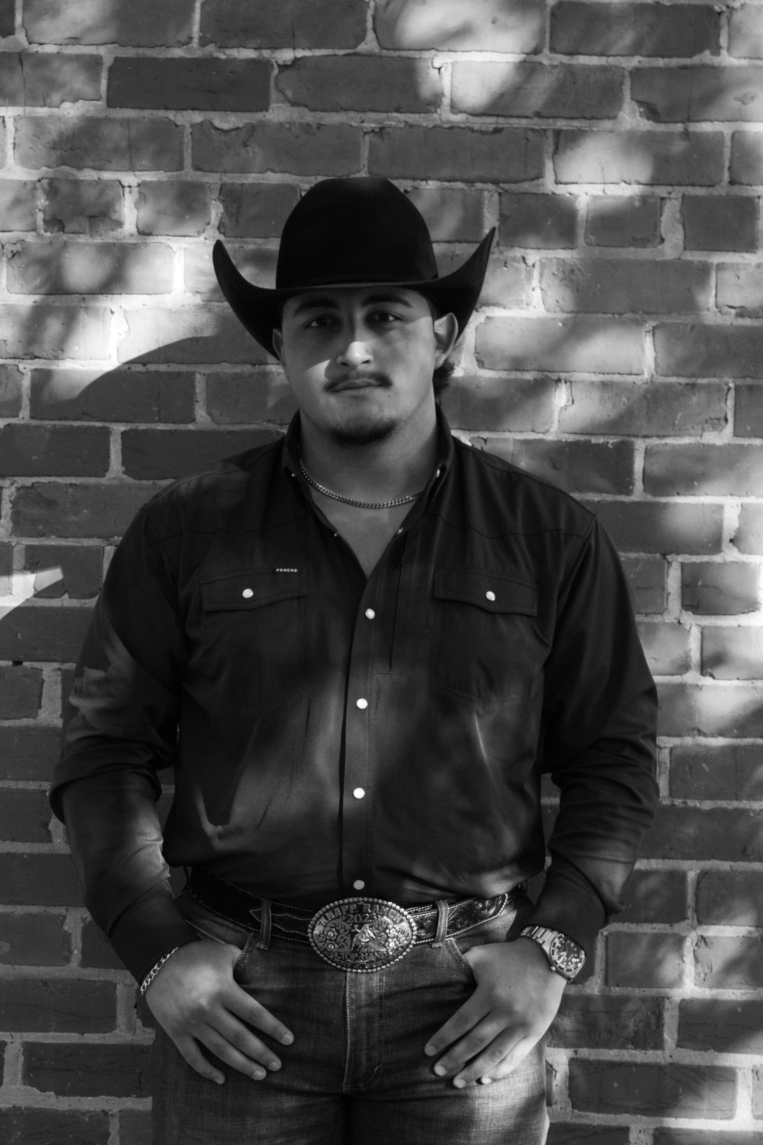 A person in western wear sits against a textured brick wall with dramatic lighting.