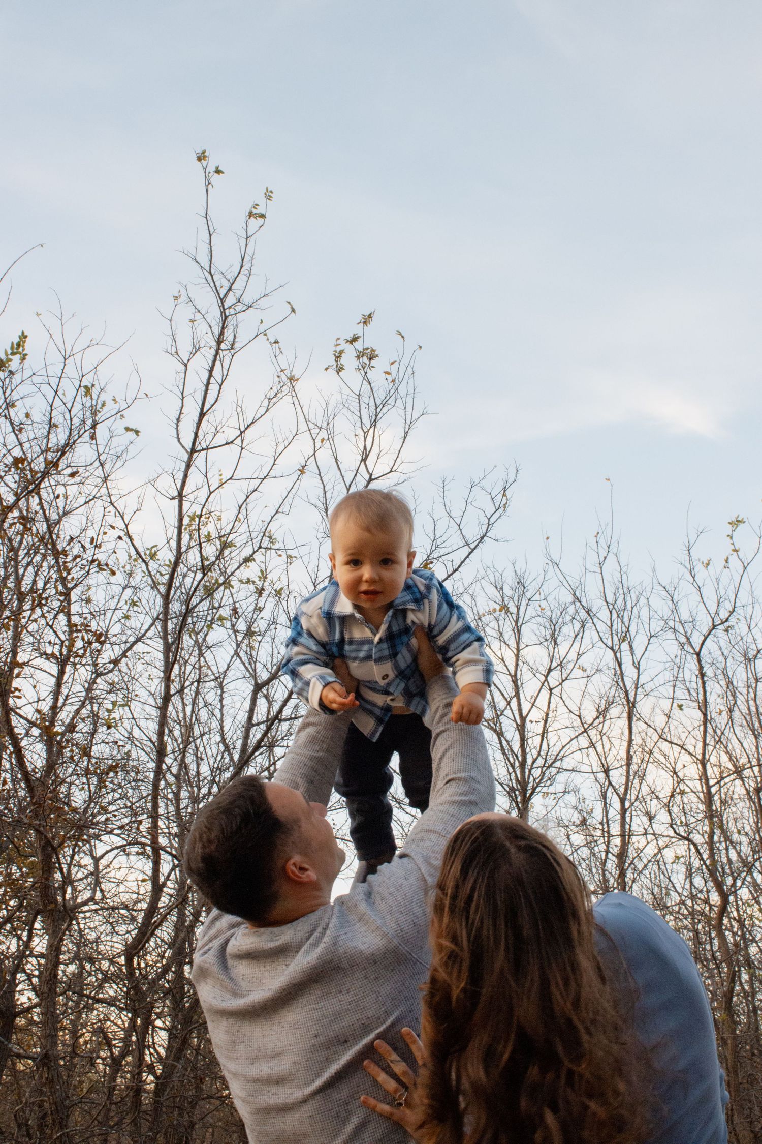 A baby being lifted up into the air against a backdrop of bare winter trees and blue sky.