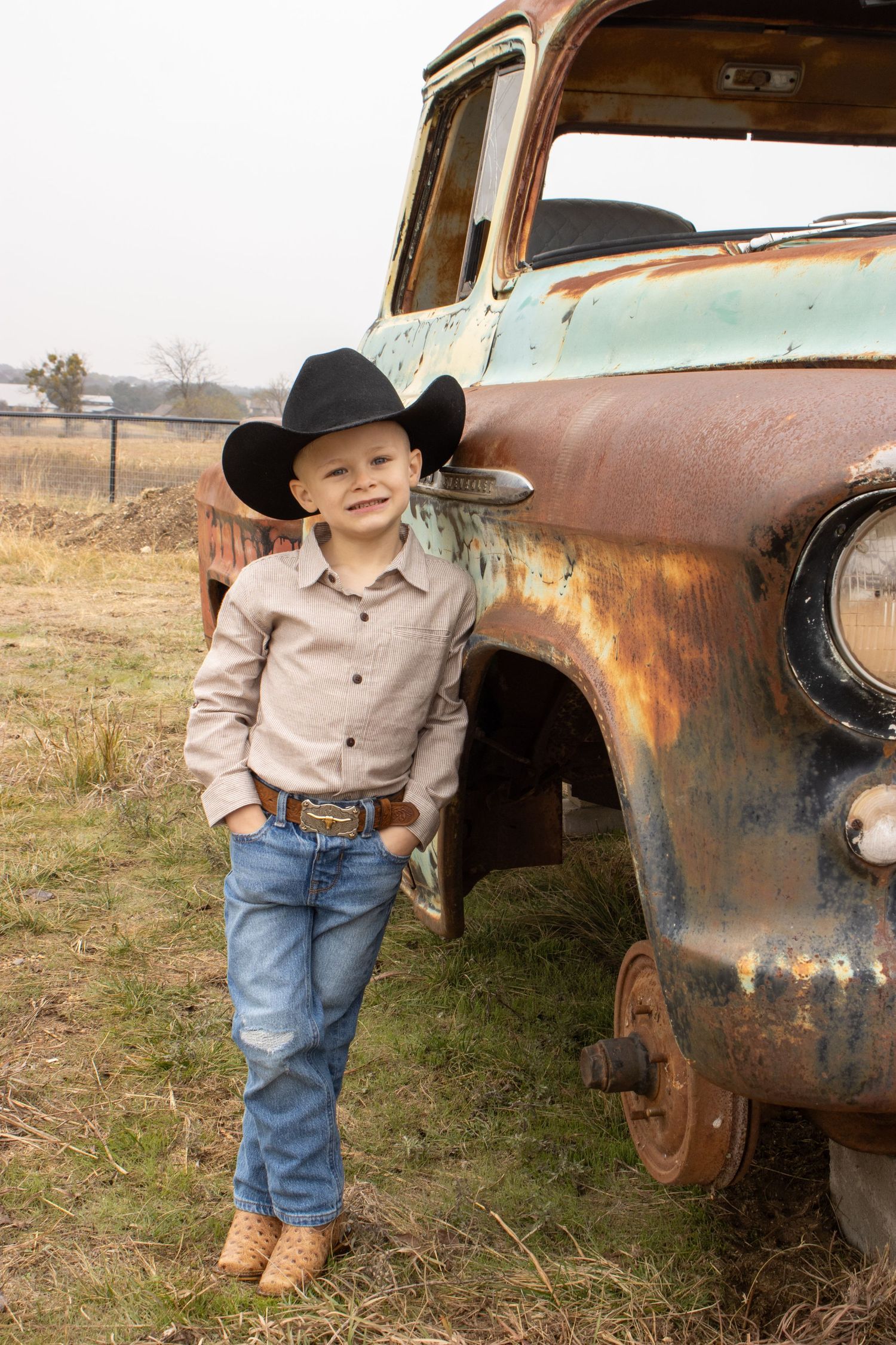 Small child in western attire poses next to rustic vintage truck in rural setting.