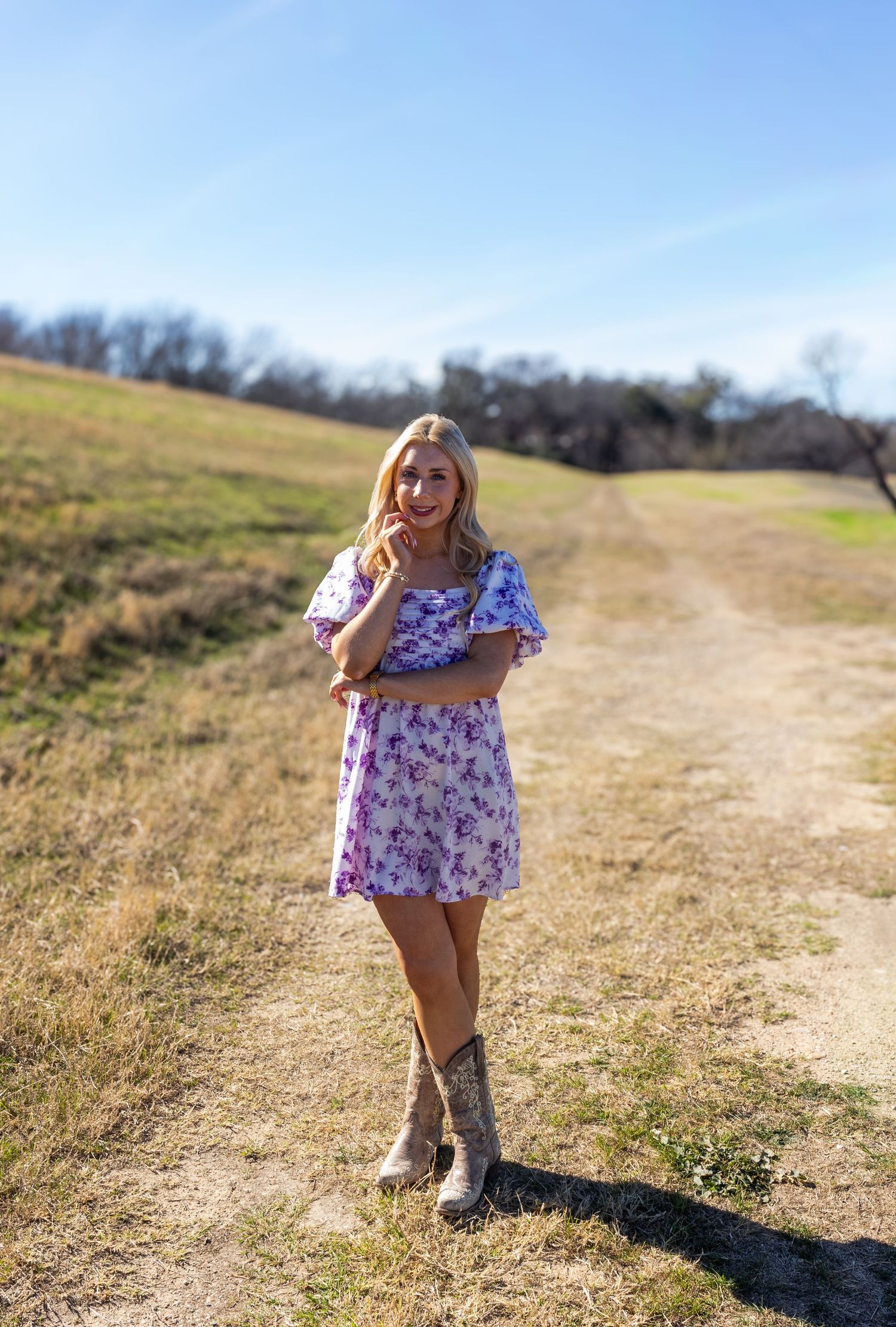 A person wearing a floral dress and boots walks along a dirt path on a sunny day in a grassy field.