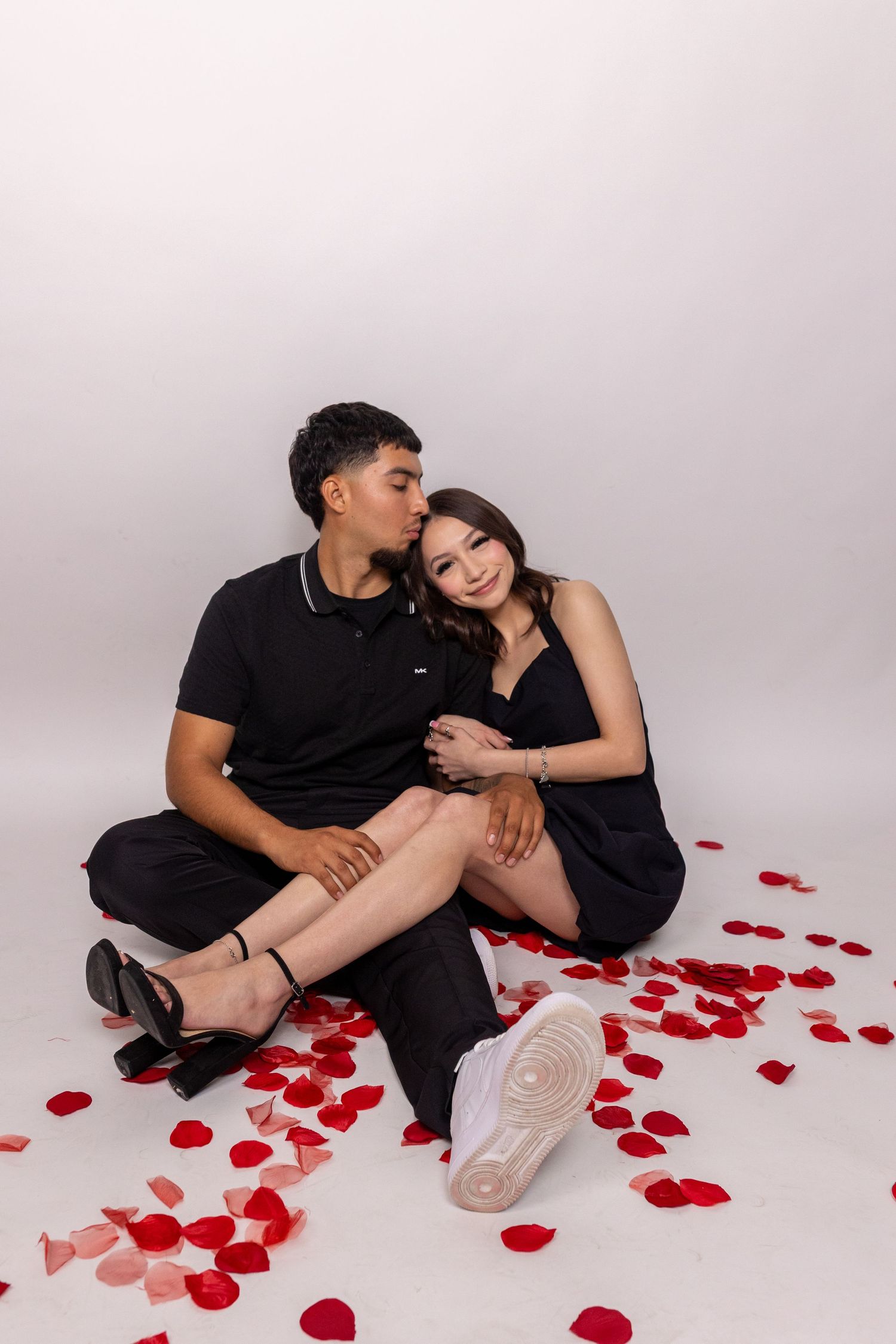 A couple in black outfits sits together on white floor with scattered red rose petals in romantic photoshoot series.