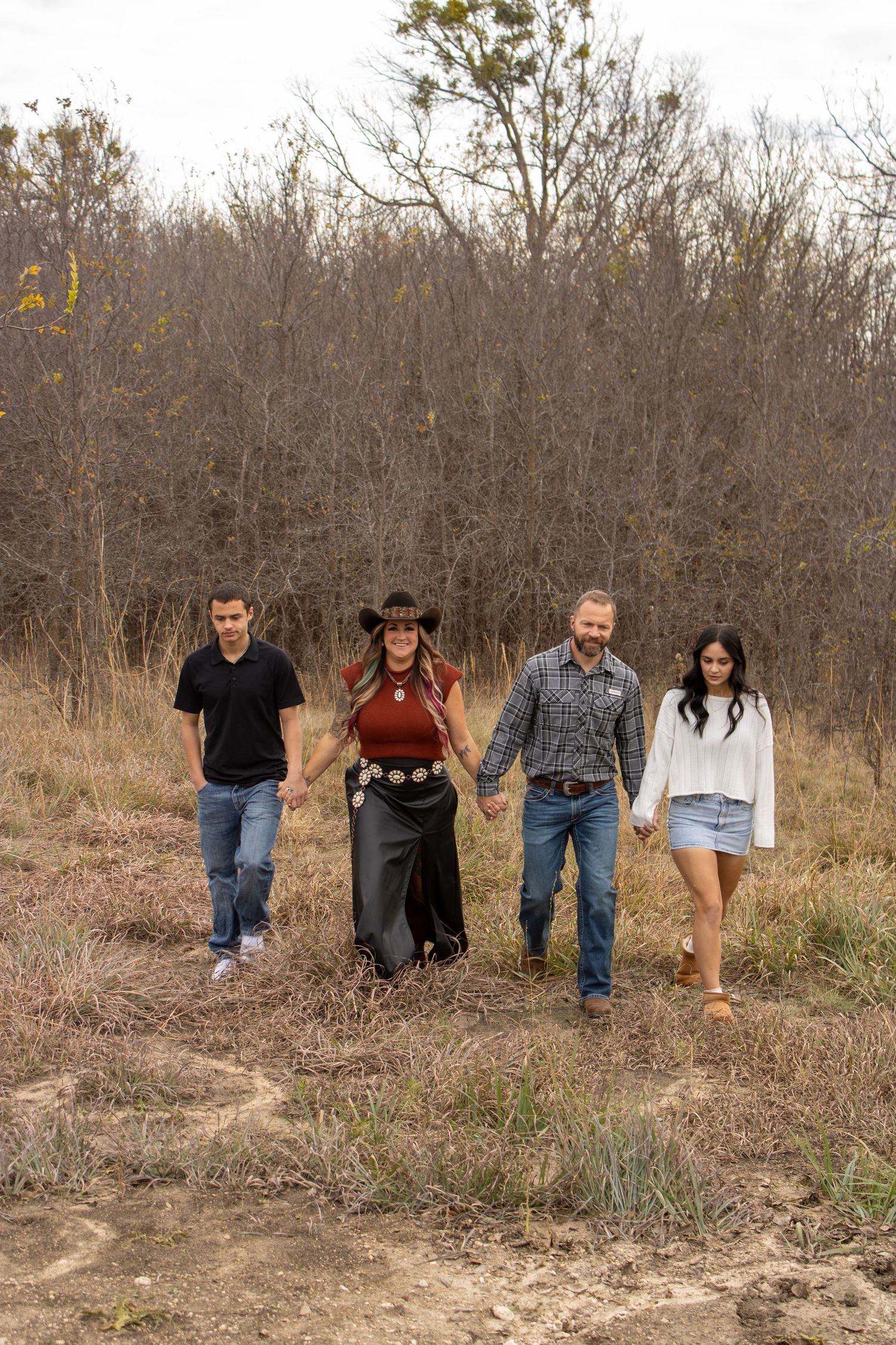 Family of four walking hand-in-hand through a rustic field, dressed in casual western attire, with bare trees in the background.