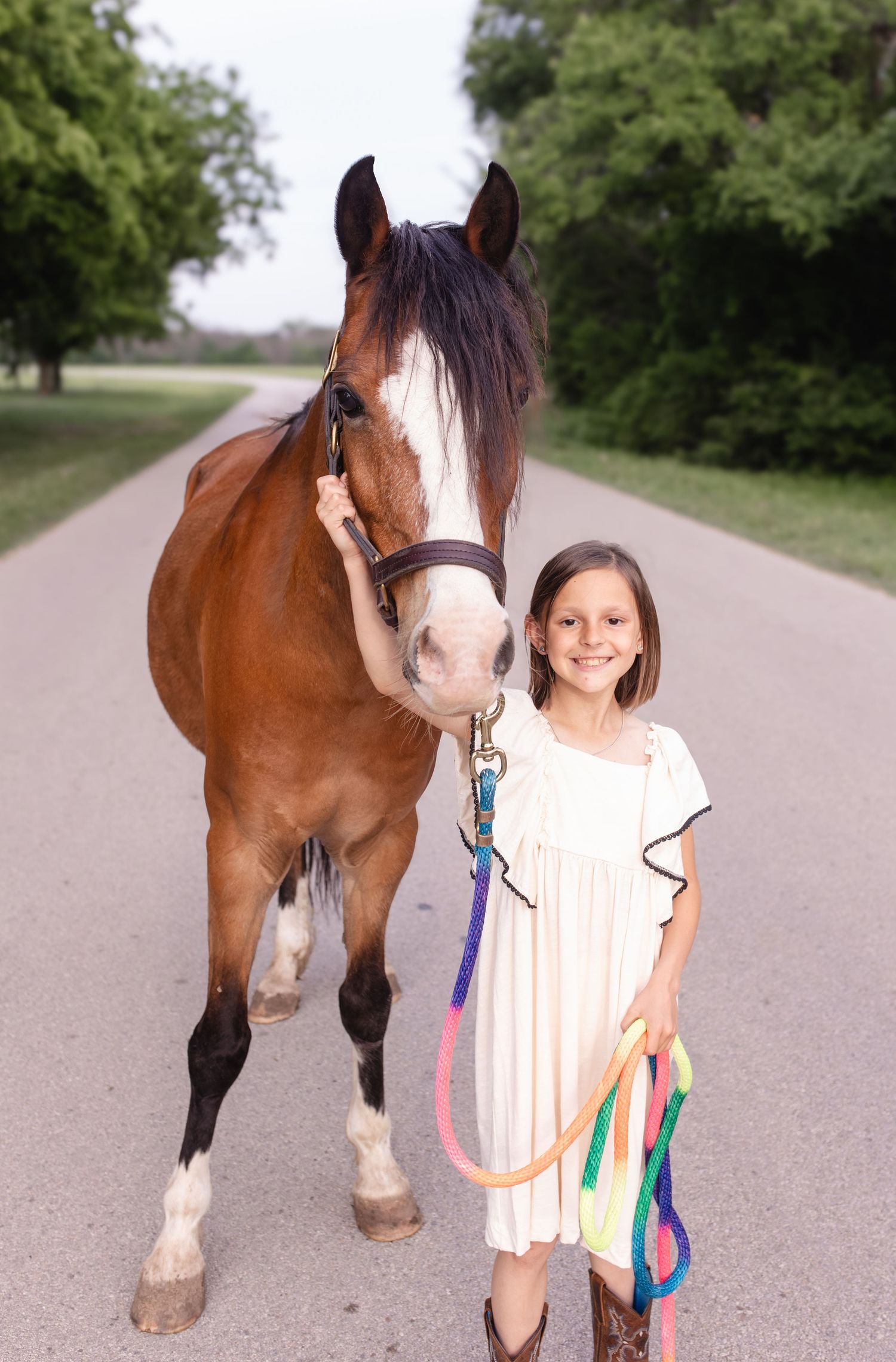 Young girl in white dress and cowboy boots smiles beside a bay horse on a tree-lined road.