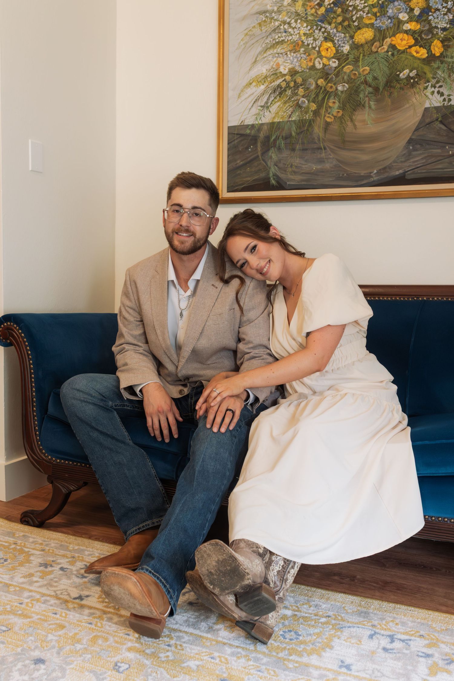 Couple sitting together on a blue velvet sofa, man in blazer and jeans, woman in white dress with cowboy boots.