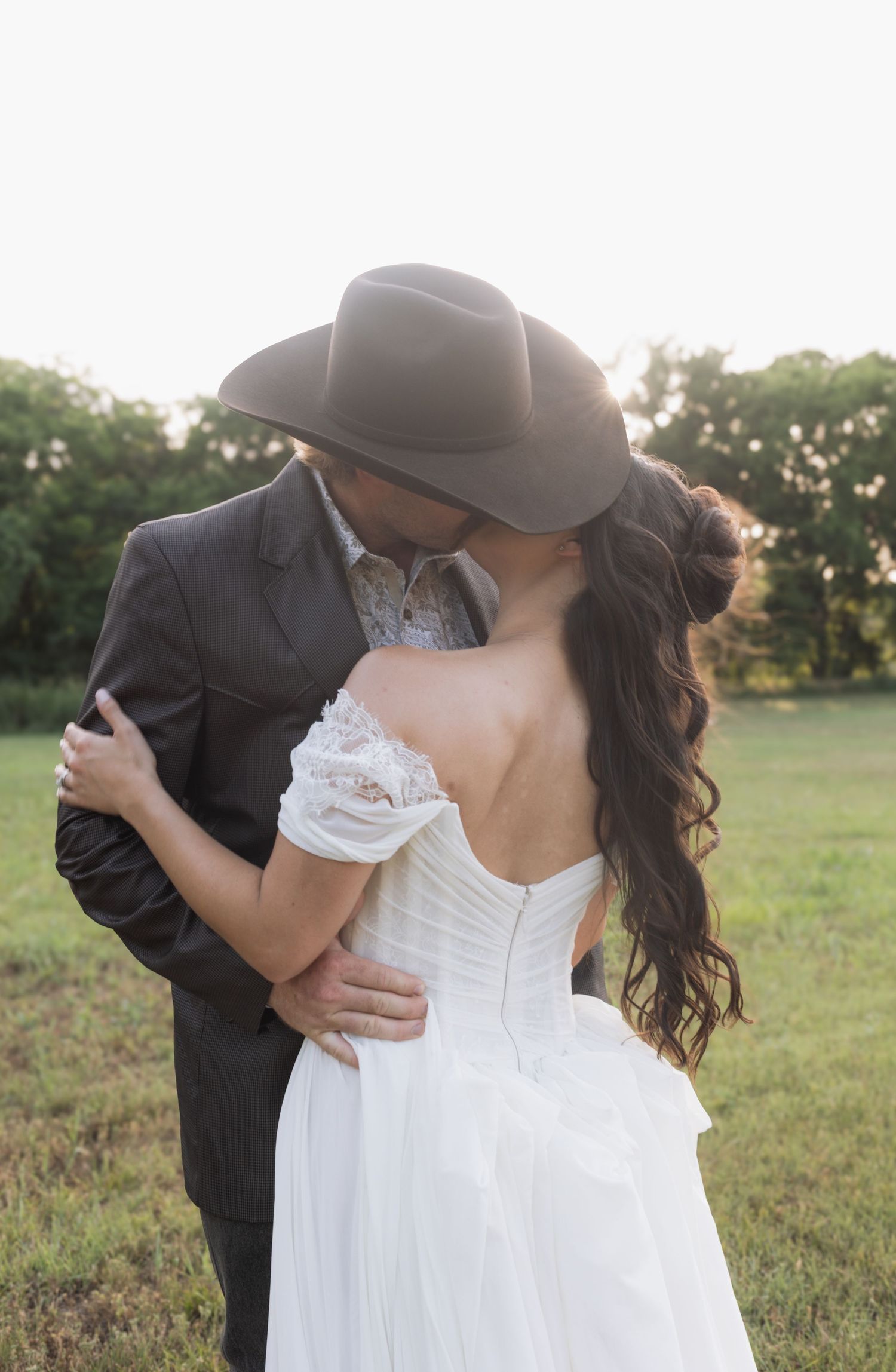 A couple shares a romantic kiss in a field, the groom wearing a cowboy hat and suit, bride in a white off-shoulder gown.
