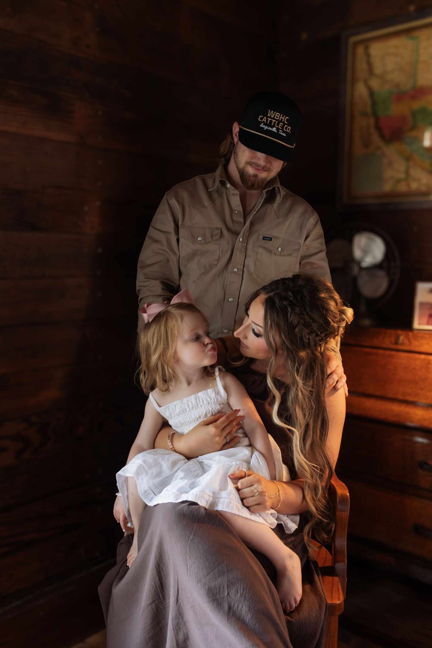 A family portrait of a man in a cap, a woman, and their toddler daughter in a rustic wooden interior.