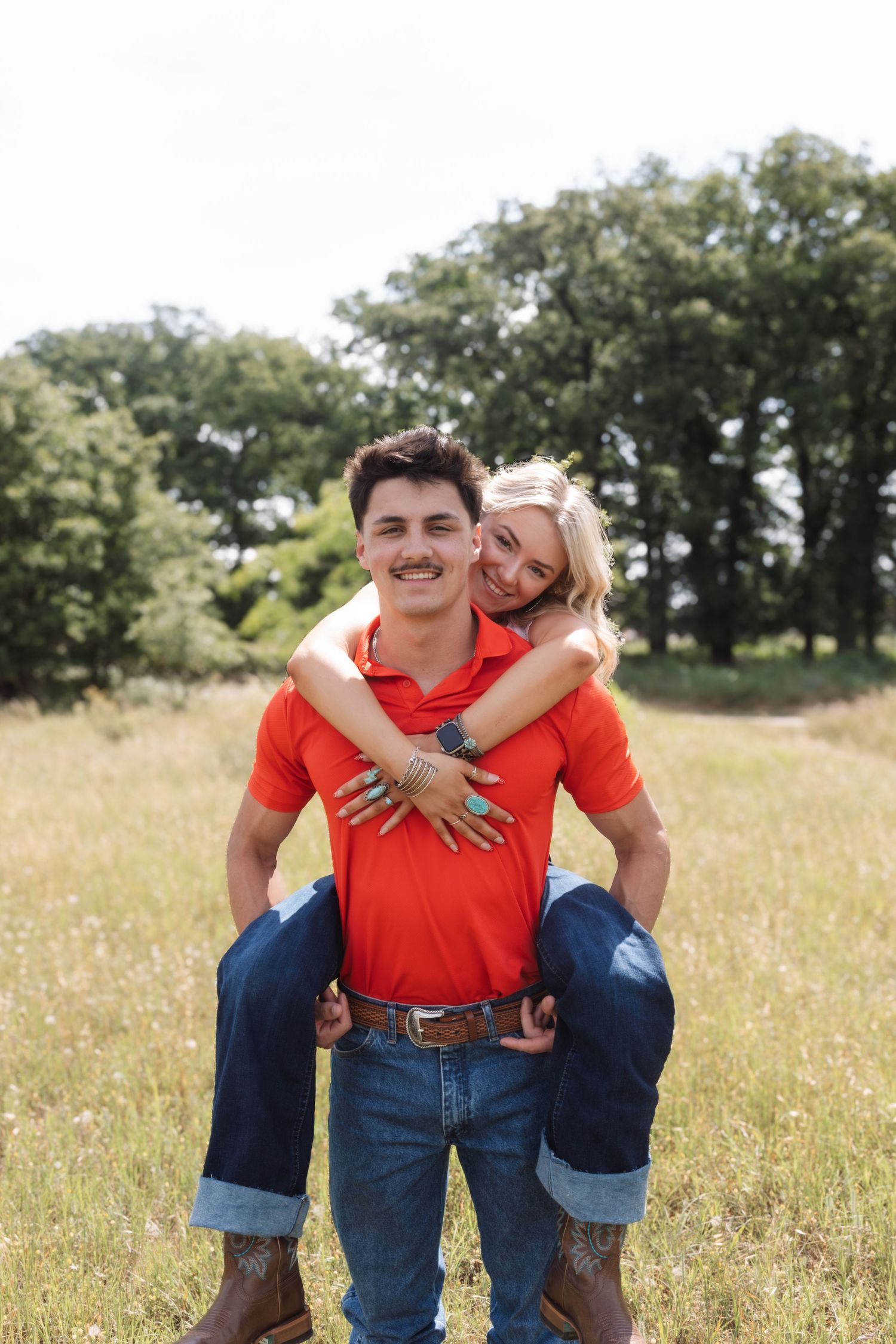 A young couple in matching red shirts and jeans share a piggyback ride in a sunny green field.