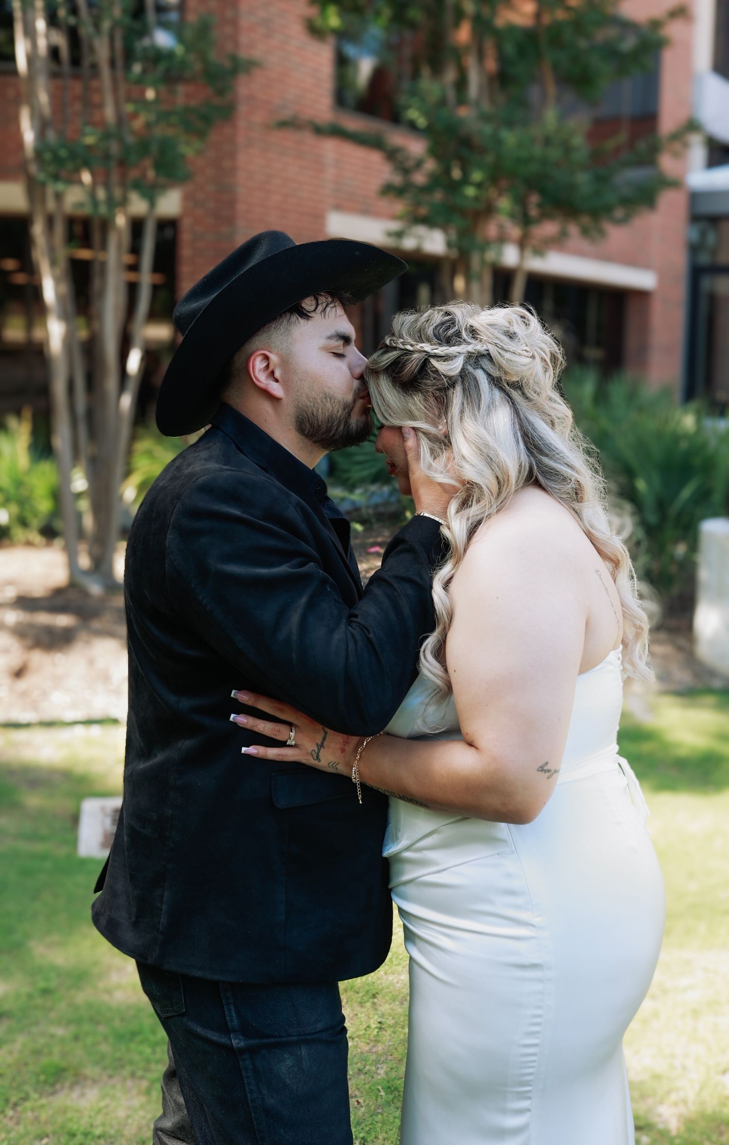Couple sharing a kiss outdoors, groom in black cowboy hat and jacket, bride in white sleeveless gown with wavy blonde hair.