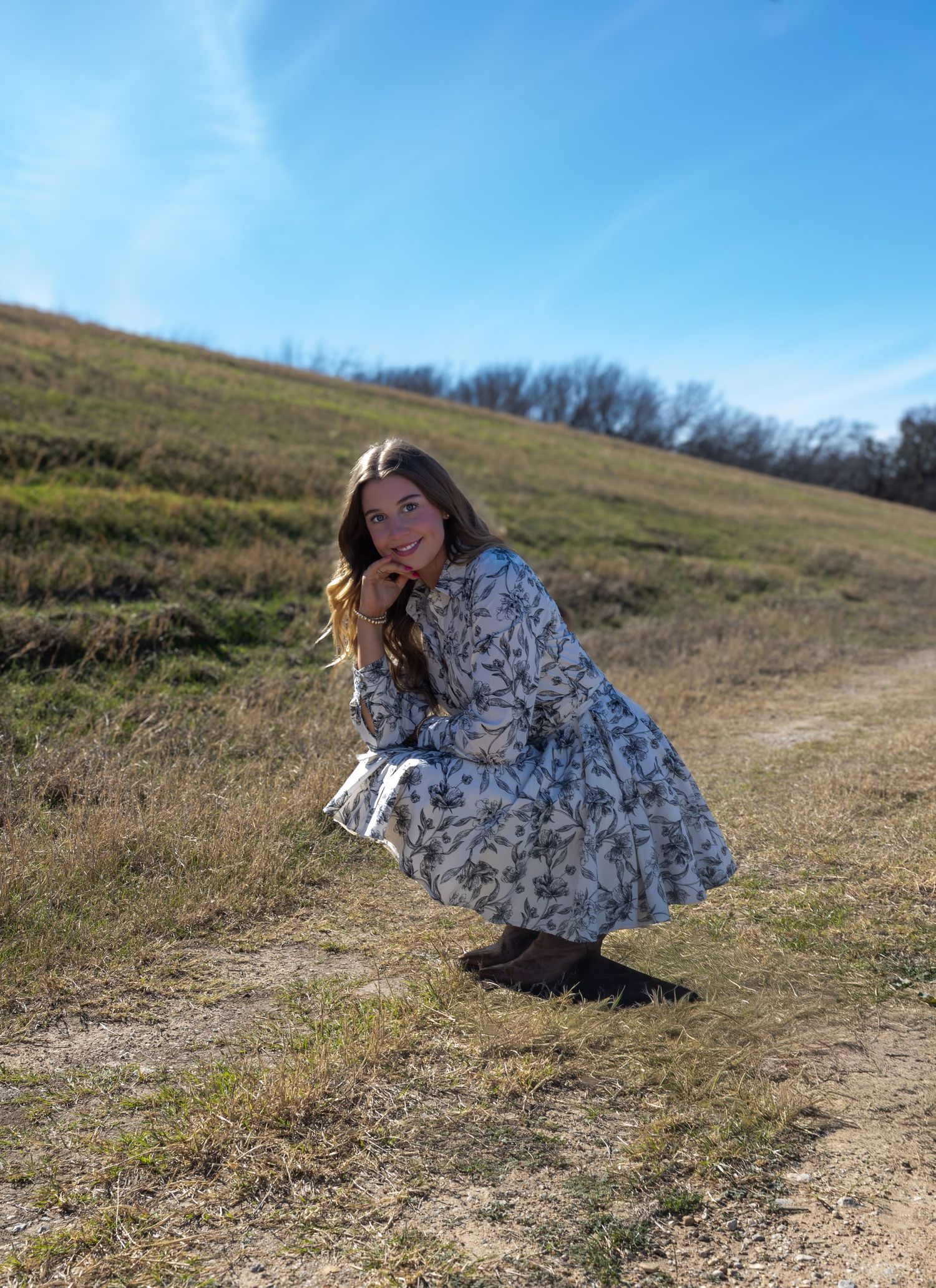 A person in a floral dress crouches down on a grassy hillside path against a bright blue sky on a sunny day.