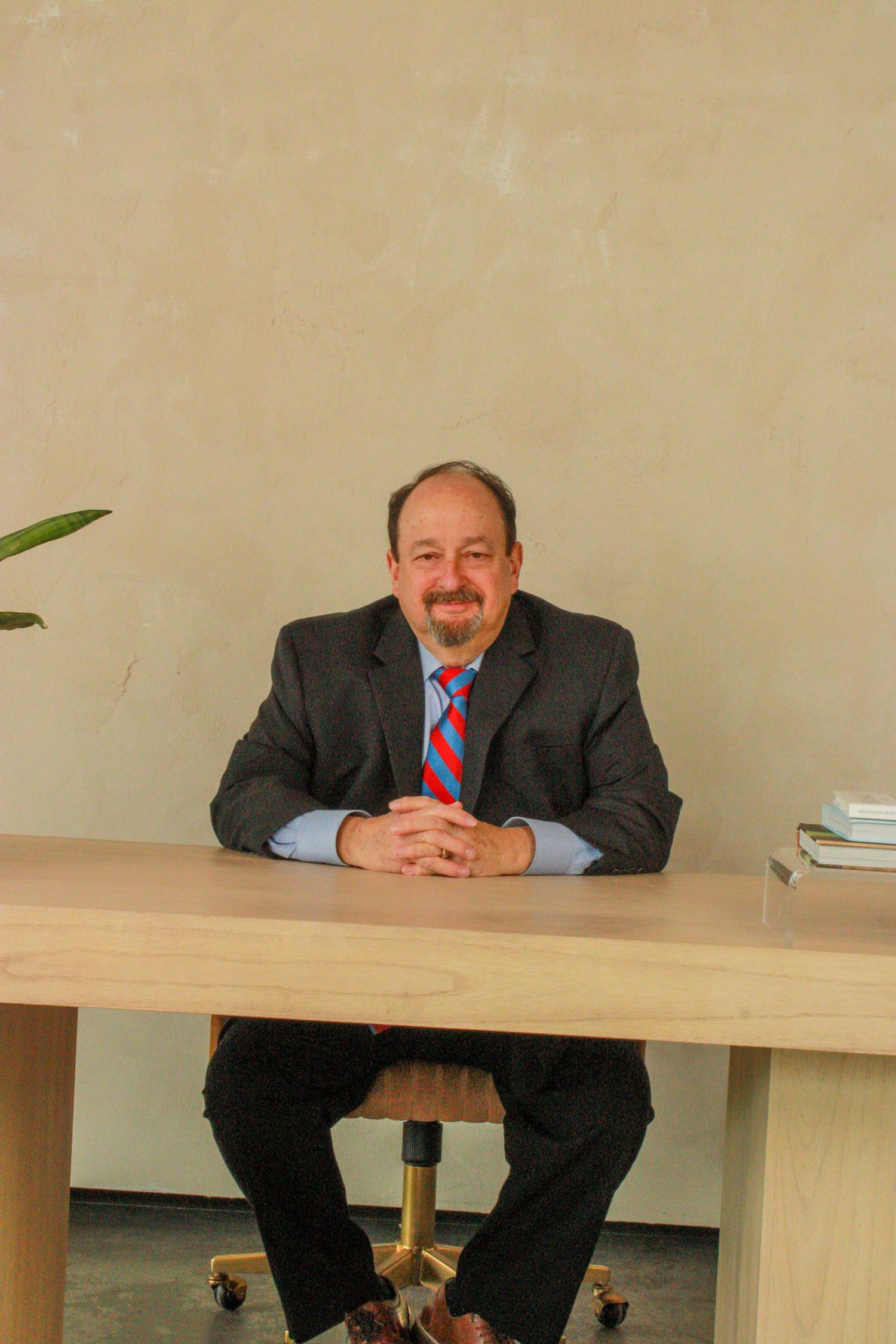 A person in a dark business suit and red striped tie sits at a light wooden desk with hands clasped.