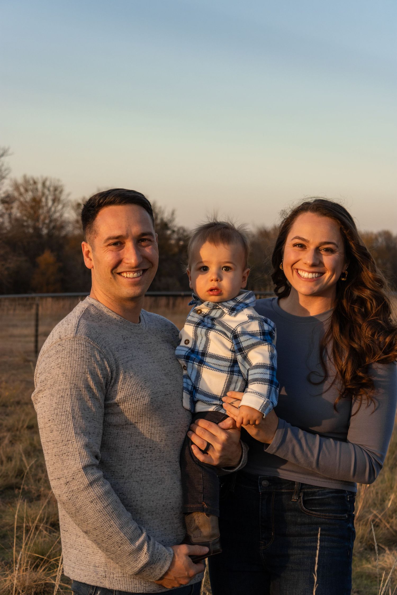 A family of three poses together outdoors during golden hour in a natural field setting.