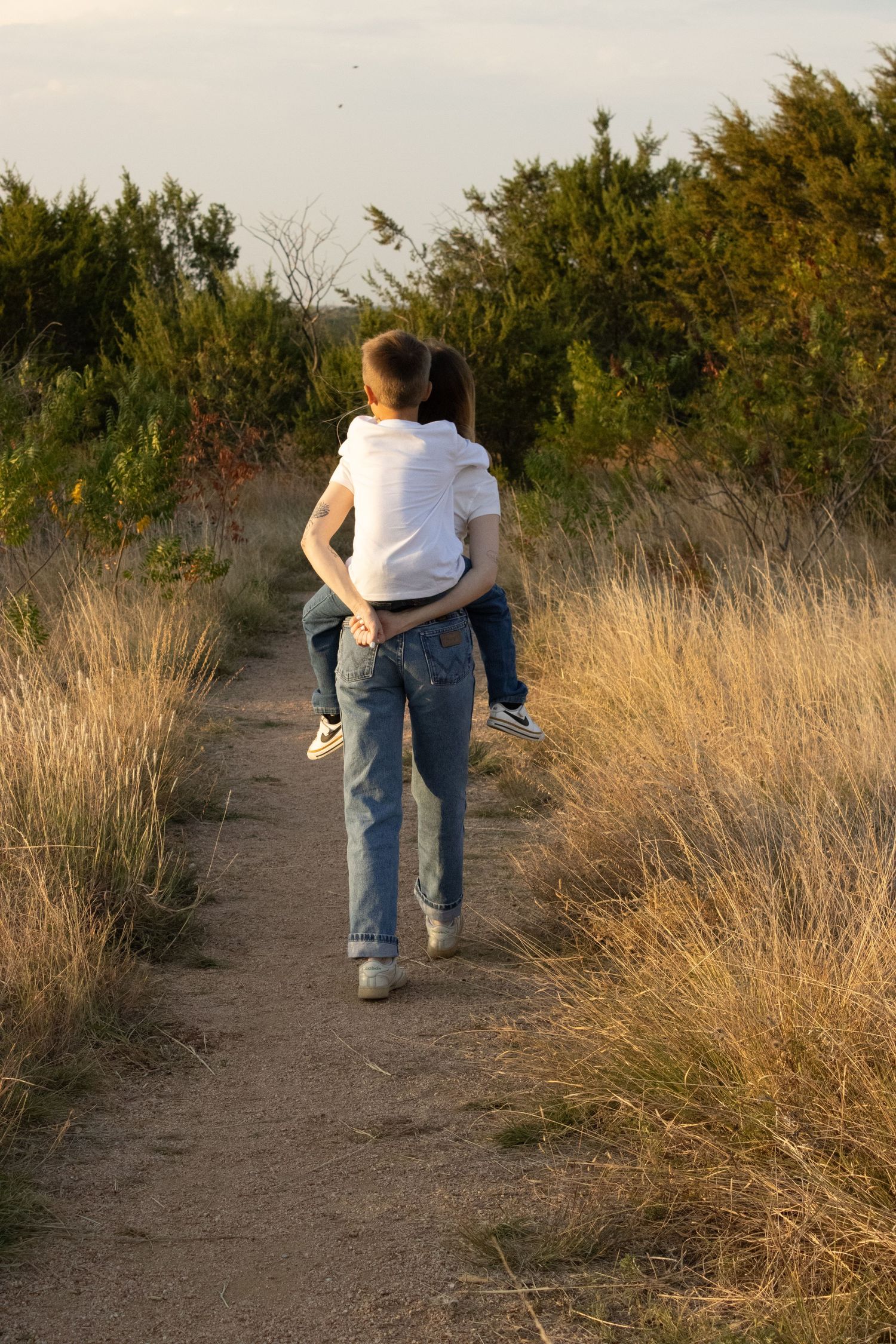 A sunset walk along a dirt path through tall golden grass and evergreen trees.