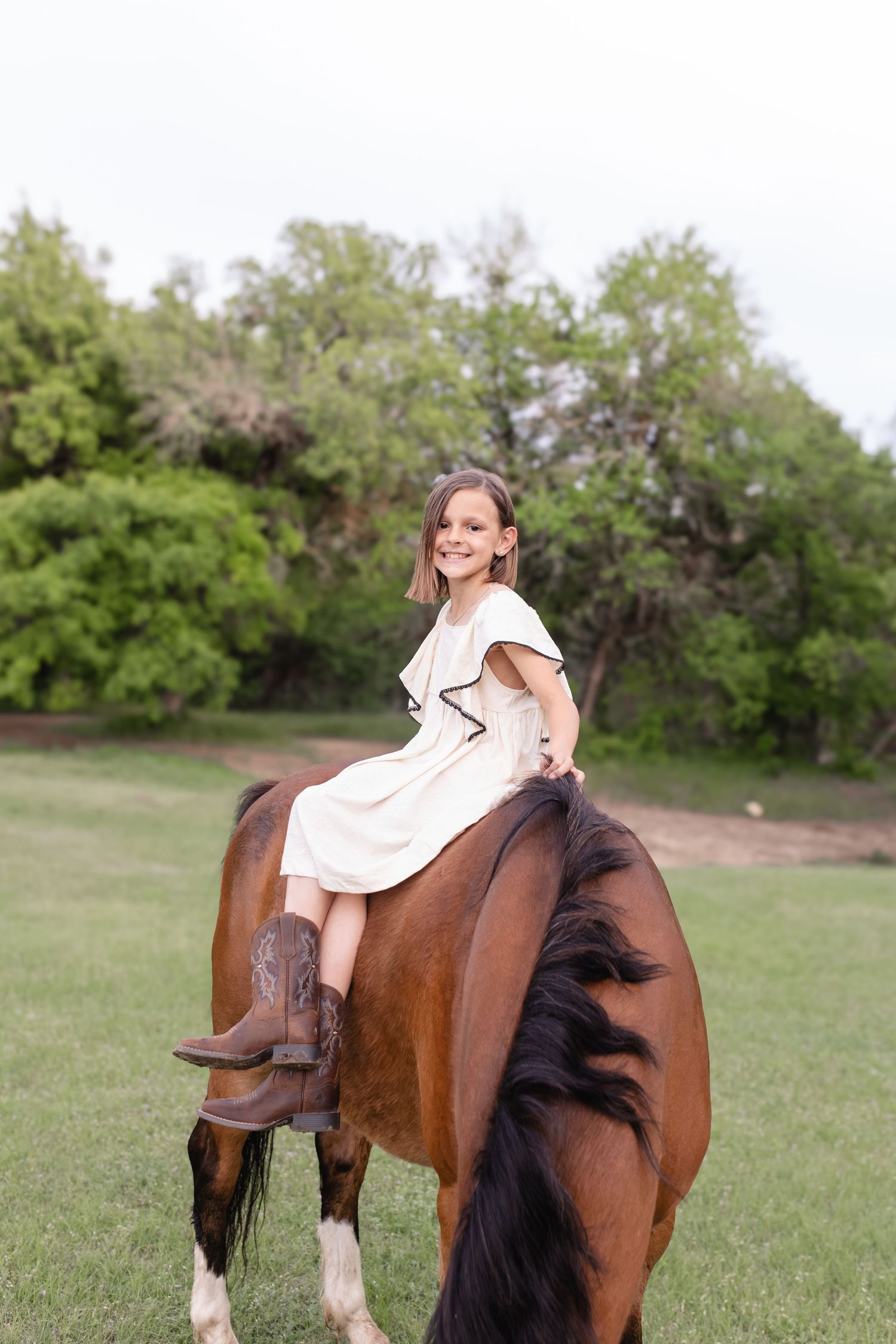 A smiling girl in a white dress and cowboy boots rides a brown horse in a lush green field.