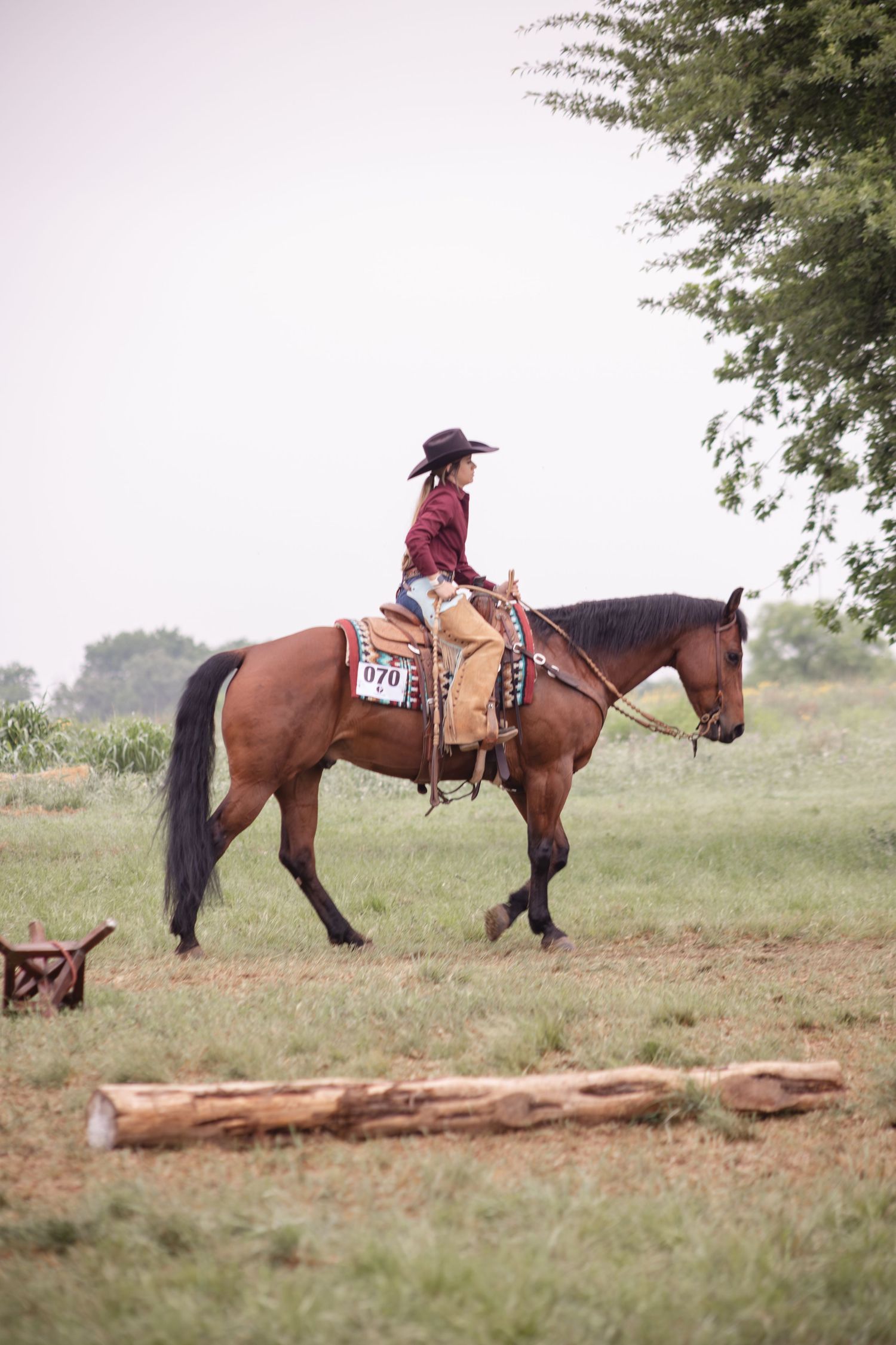 A cowgirl in a red shirt and black hat rides a bay horse numbered 070 in an outdoor equestrian competition.