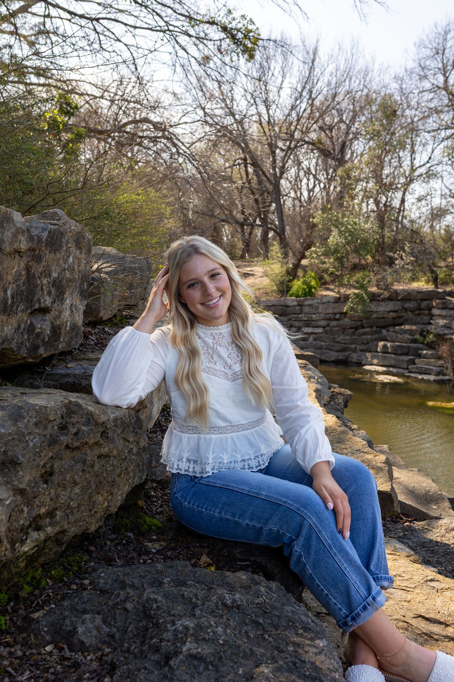 A person in a white sweater and jeans sits on rocky outcrops by a pond on a sunny winter day.
