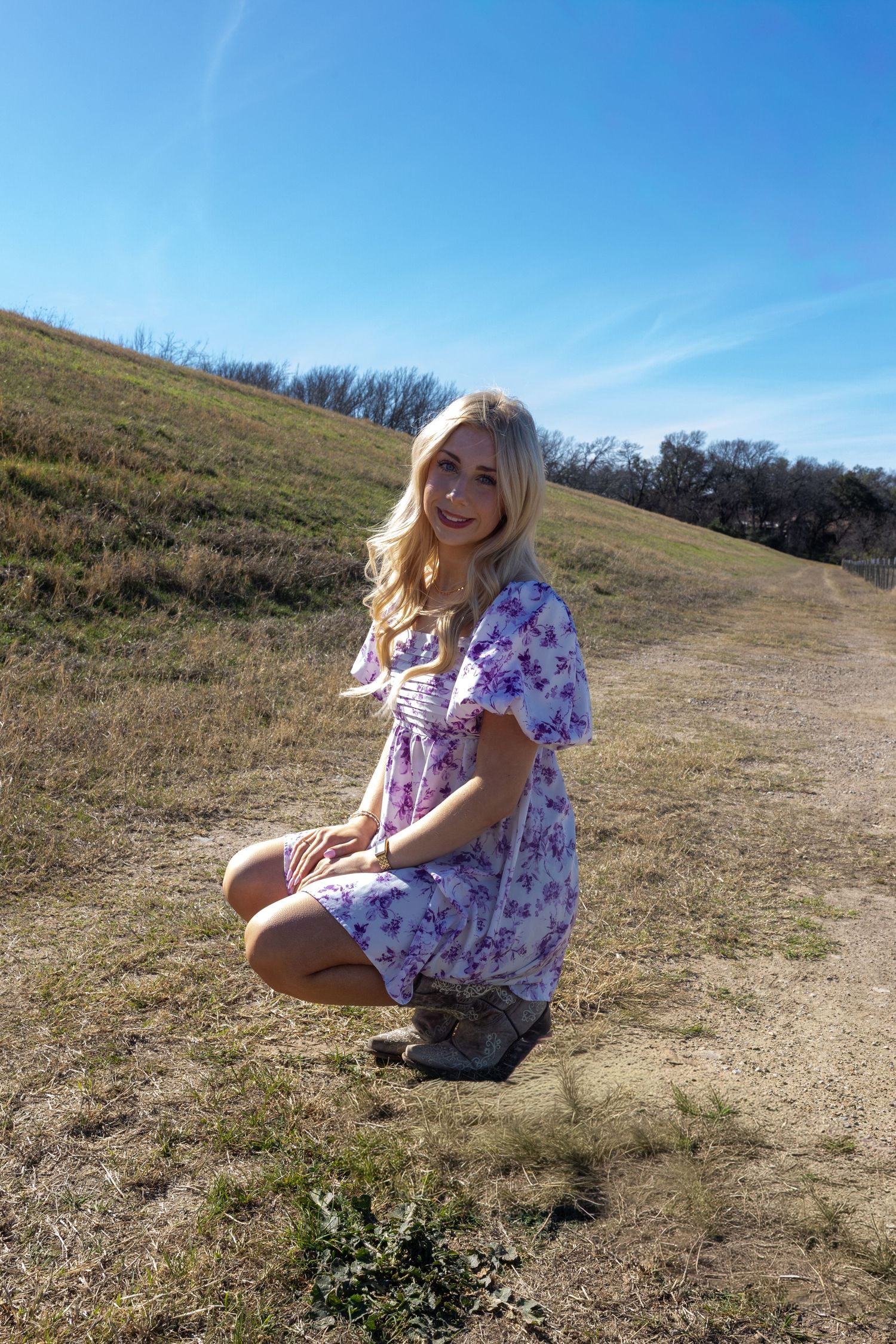 Someone in a purple floral dress and boots crouches on a grassy hillside trail on a sunny day with blue skies.