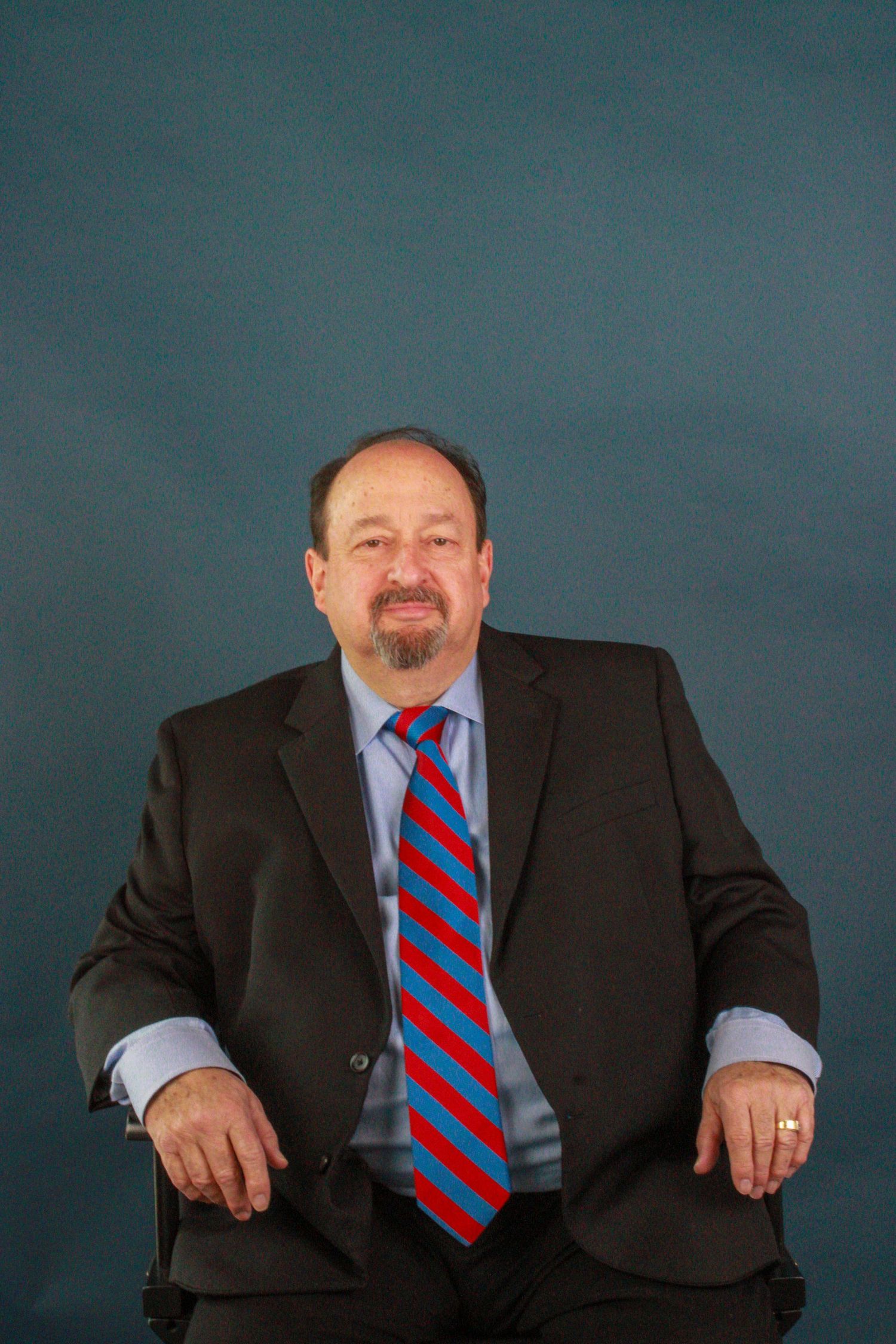 Professional portrait against blue backdrop showing person in dark suit with red and navy striped tie.