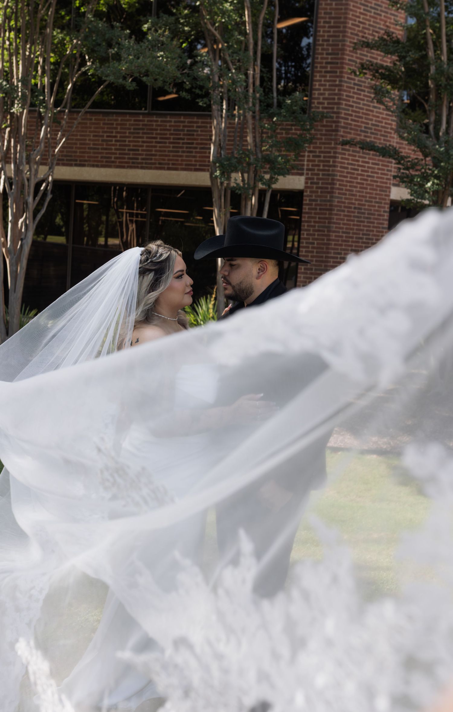 Bride in white gown with flowing veil and groom in black cowboy hat share a romantic moment outdoors near brick building.