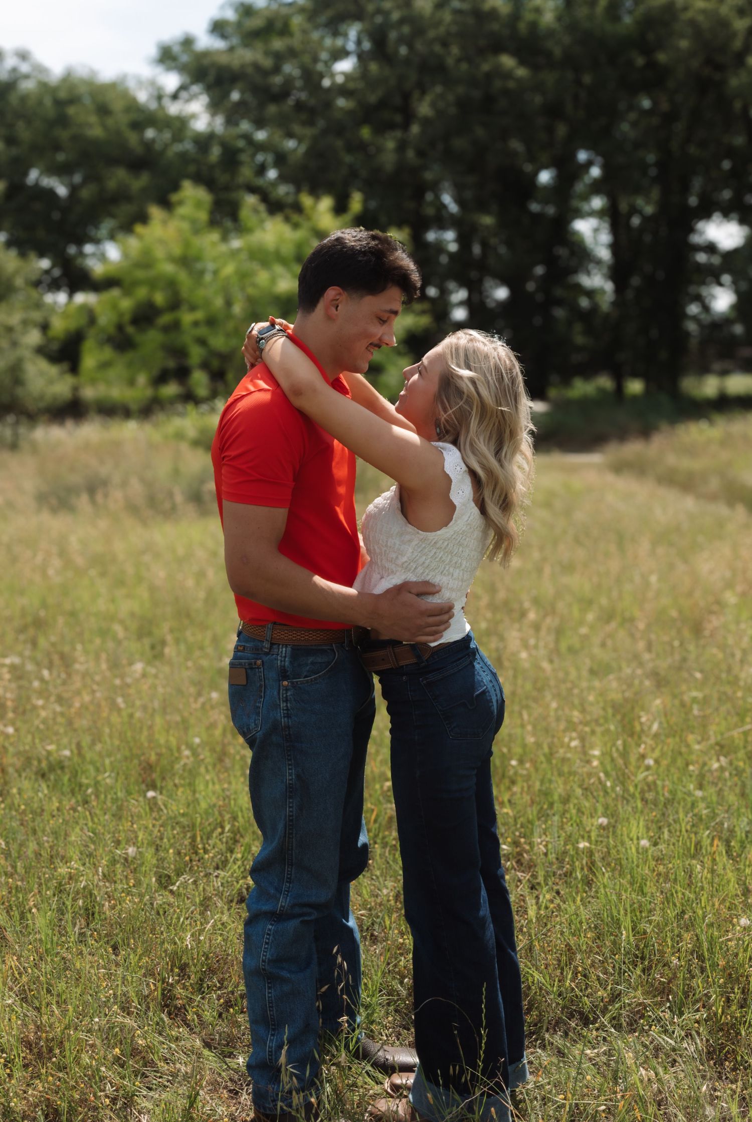 Couple embracing in a grassy field, man in red shirt and woman in white top with jeans, surrounded by green trees.