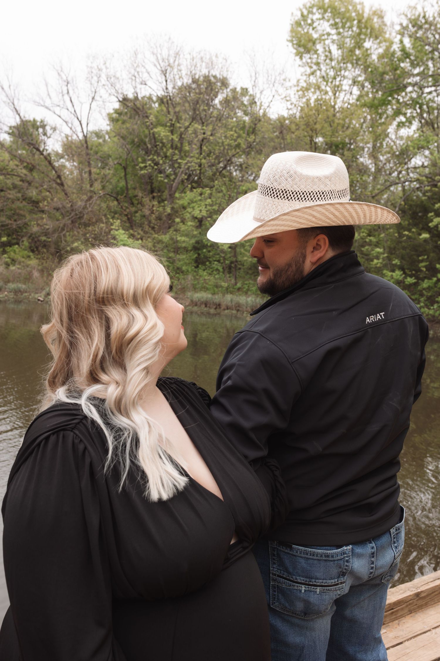 A couple in black clothing and cowboy hats share intimate moments by a lake during an outdoor photoshoot.