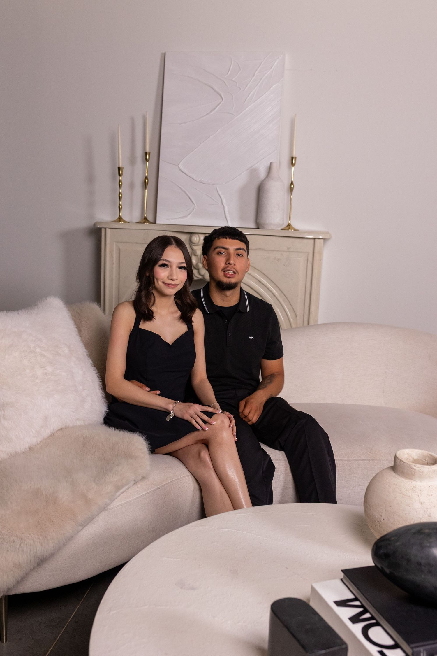 A couple in matching black outfits sits together on a white couch with candles and decor in the background.