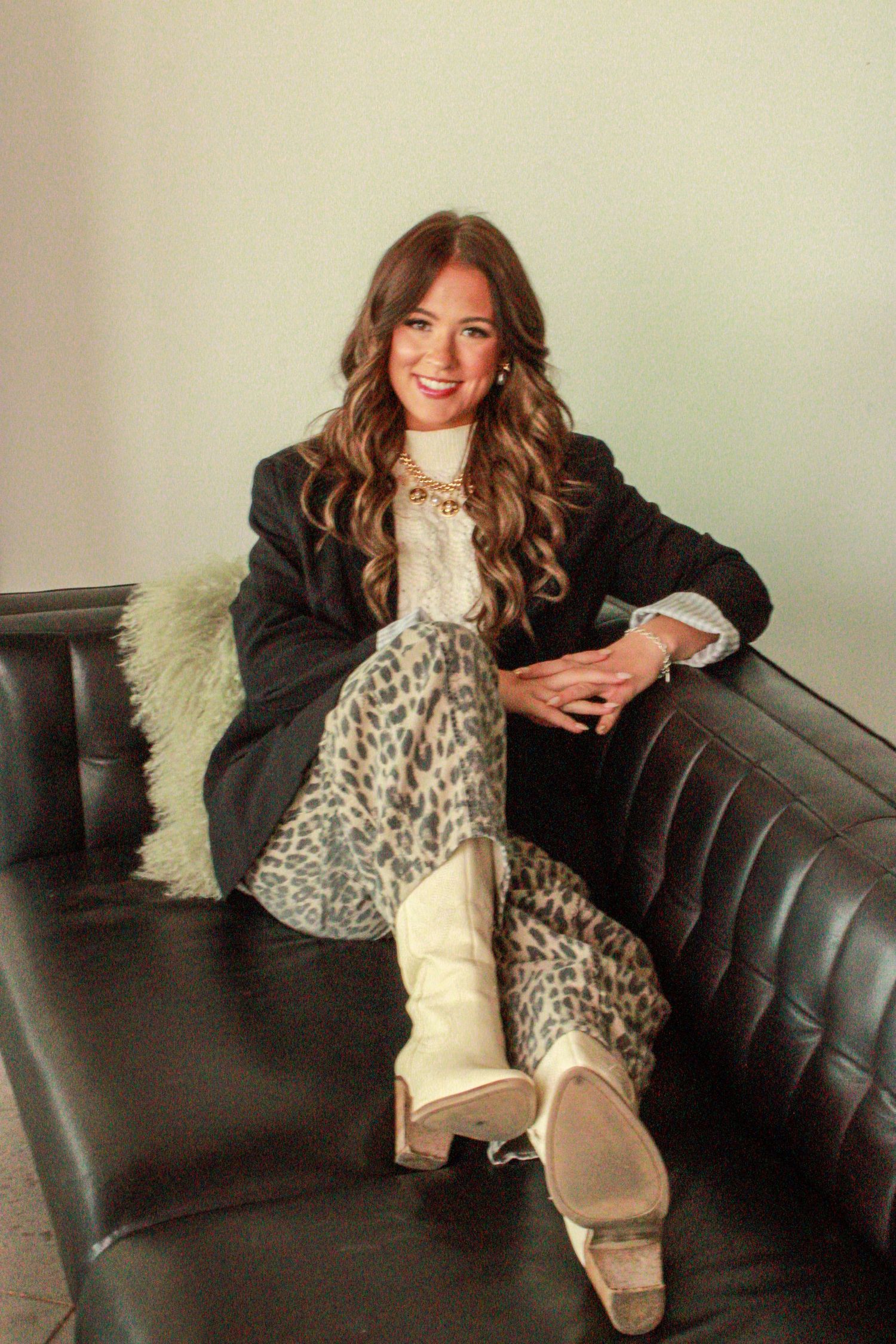 Woman relaxing on black leather couch wearing leopard print scarf and black blazer with beige boots.