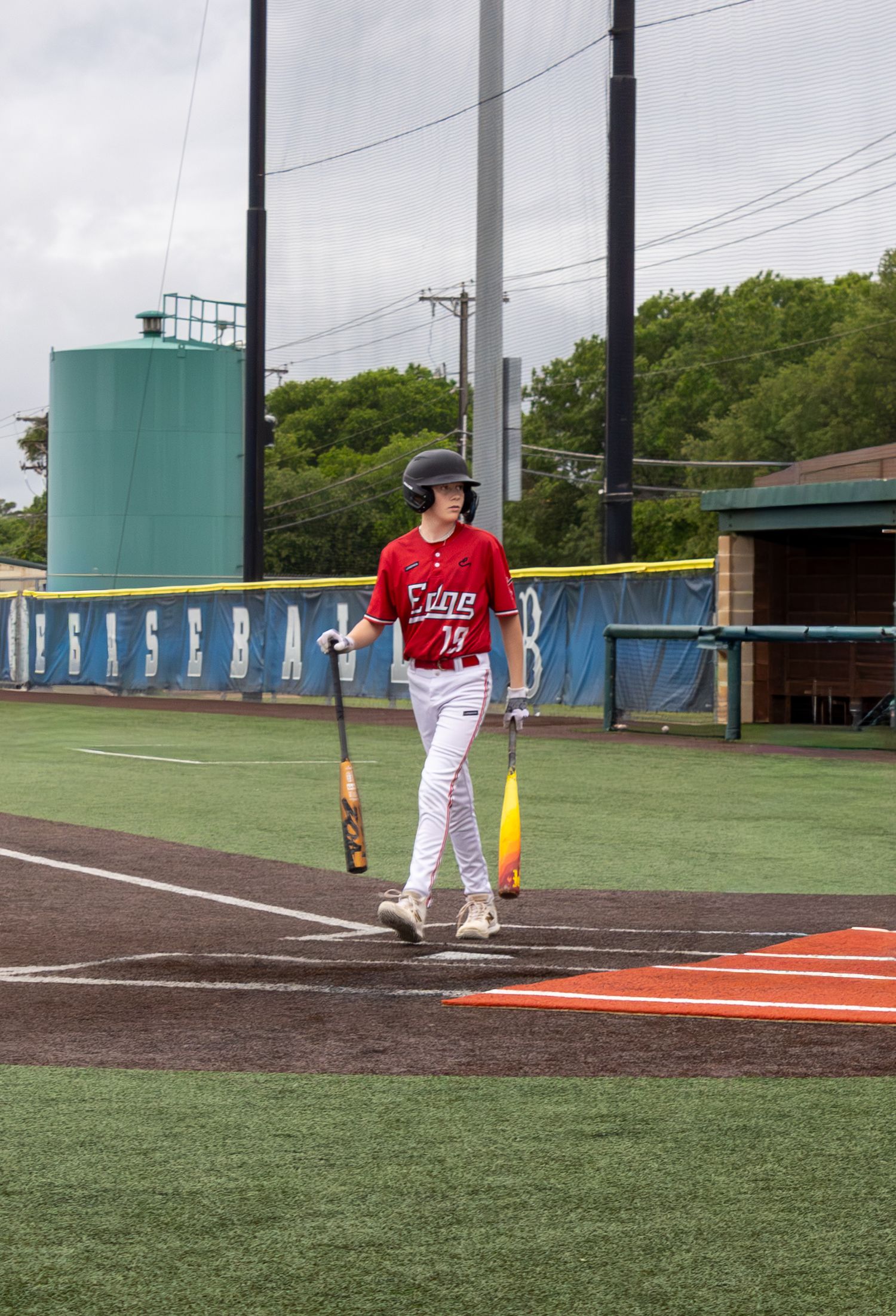 Baseball player in red jersey holding bat walks near home plate on an outdoor baseball field.