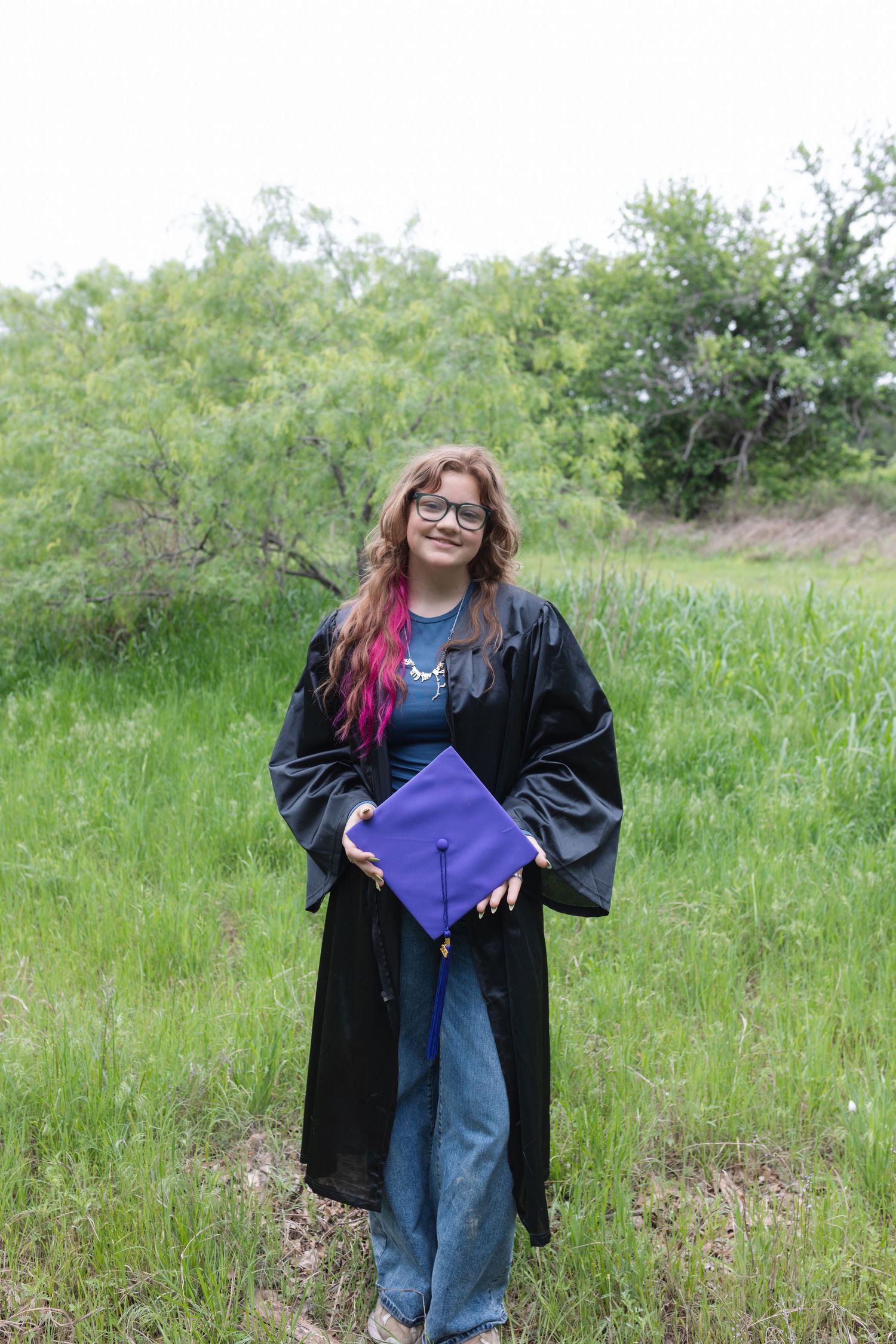 Young woman in black graduation gown holding purple mortarboard cap, standing in green field with trees behind her.