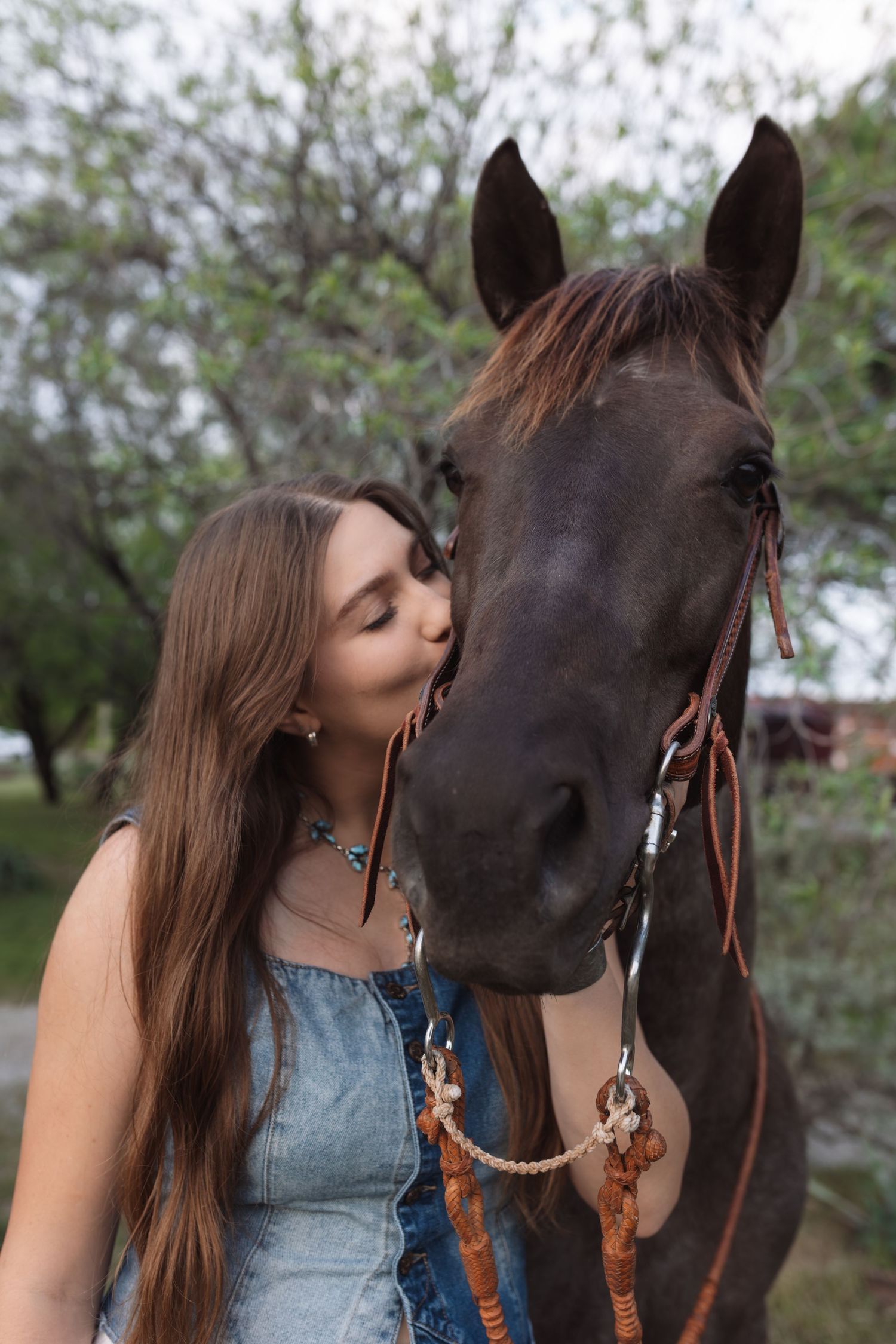 Young woman with long hair kissing a dark brown horse on the nose outdoors near trees.