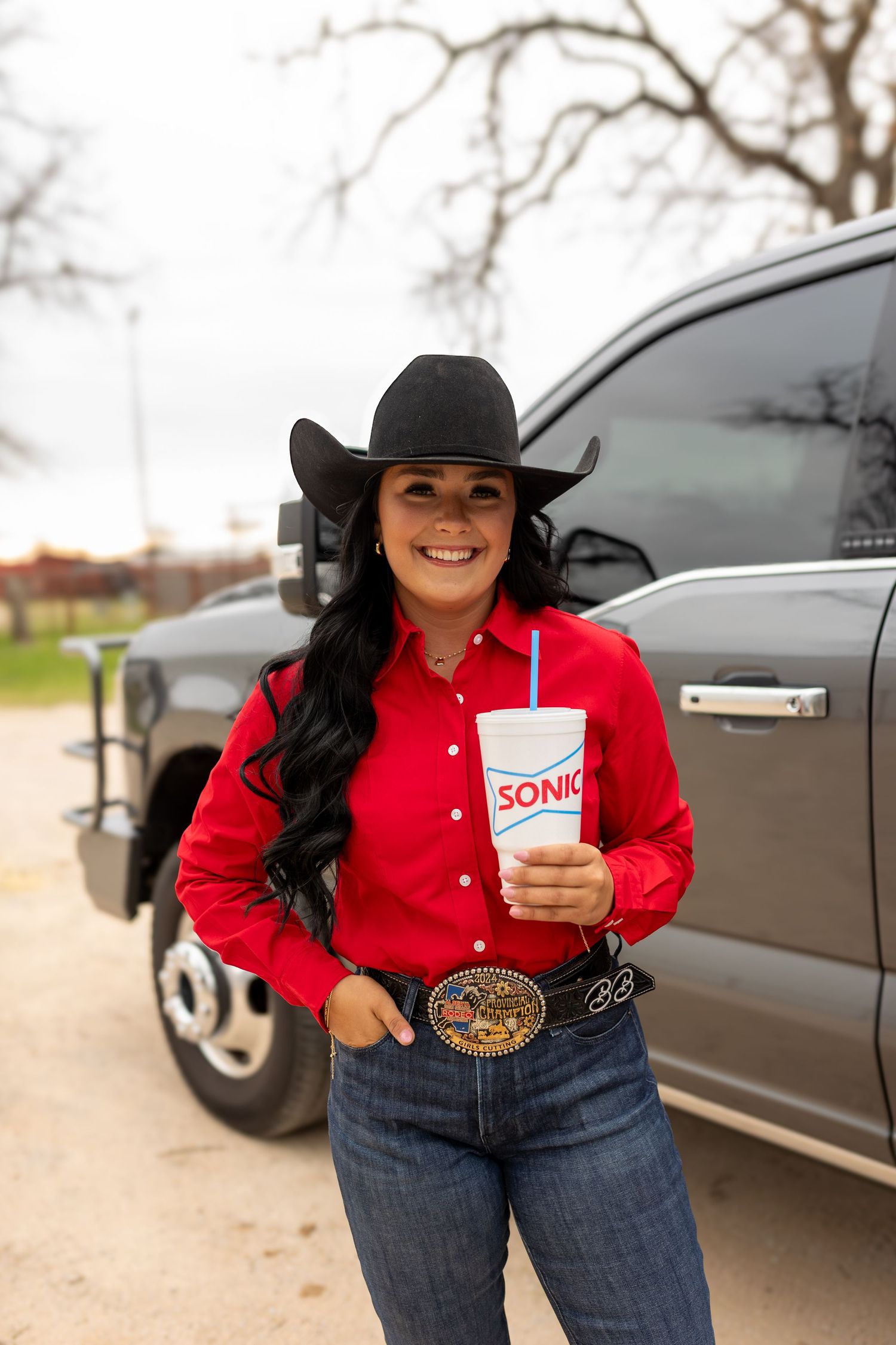 Person in red western shirt and cowboy hat holding a Sonic drink cup next to a black vehicle.